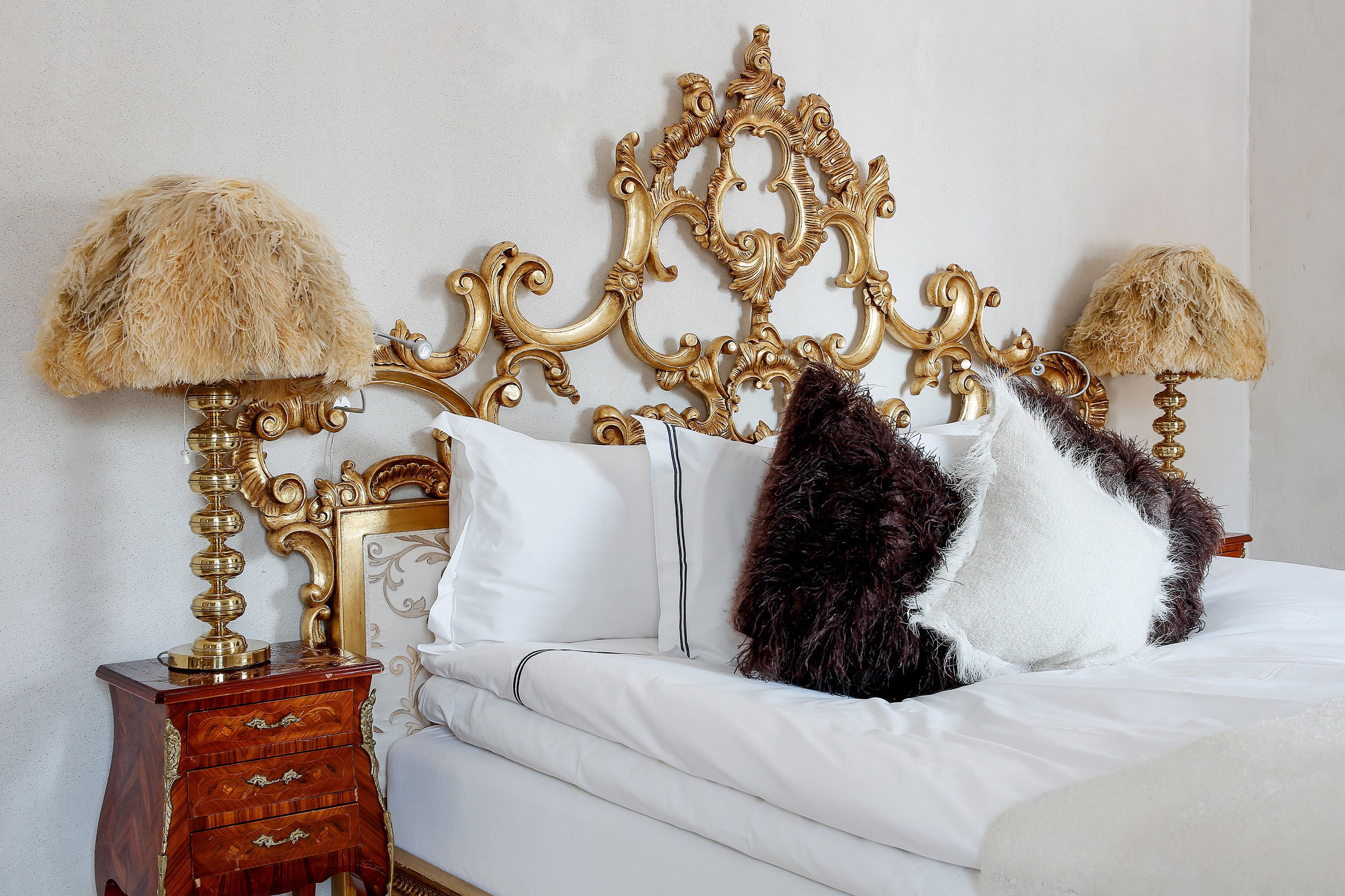 A bed with a gold ornate headboard, white bedding, and two lamps with shaggy lampshades.