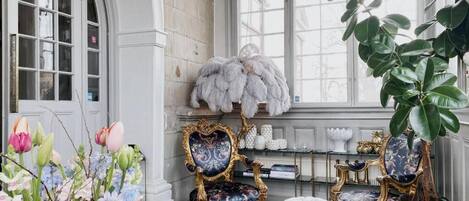 A room with a grand window, ornate chairs, a plush fur throw, and a large potted plant.