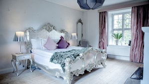 A classic four-poster bed with ornate detailing, a nightstand with a lamp, and a view of greenery through a window with curtains. - Görvälns Slott (Jarfalla)