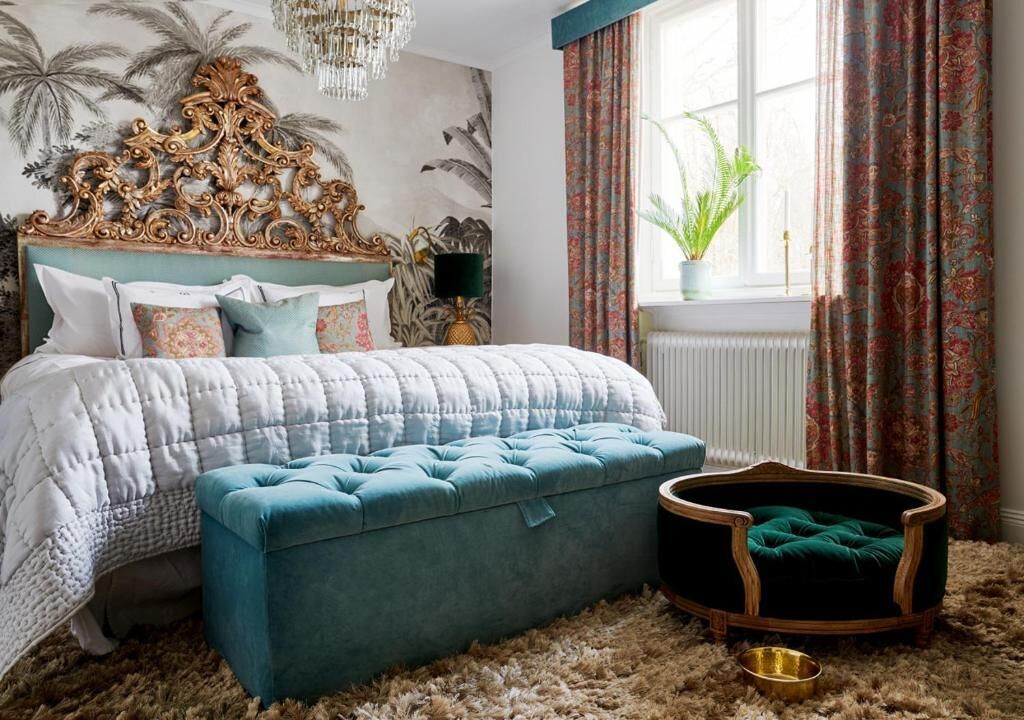 A bedroom with a bed, a tufted ottoman, a pet bed, a chandelier, and a window with curtains.