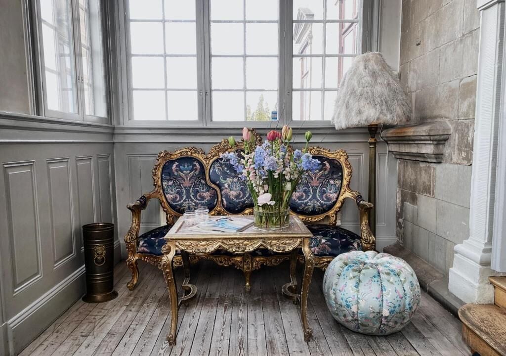 Ein Zimmer mit einem alten Sofa, einem kleinen Tisch mit Blumen in einer Vase und einer dekorativen Lampe.