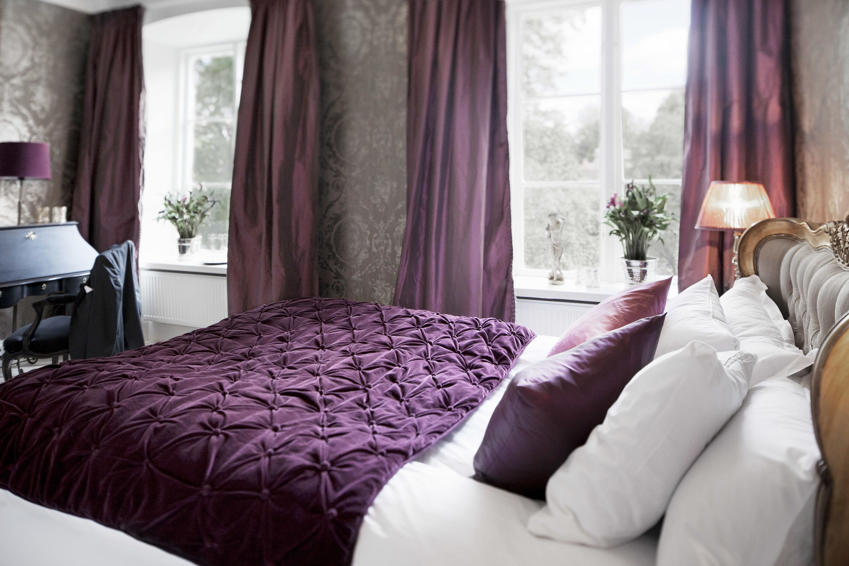 Ein Schlafzimmer mit einem großen Bett, violetten Vorhängen und einem Schreibtisch mit Lampe.