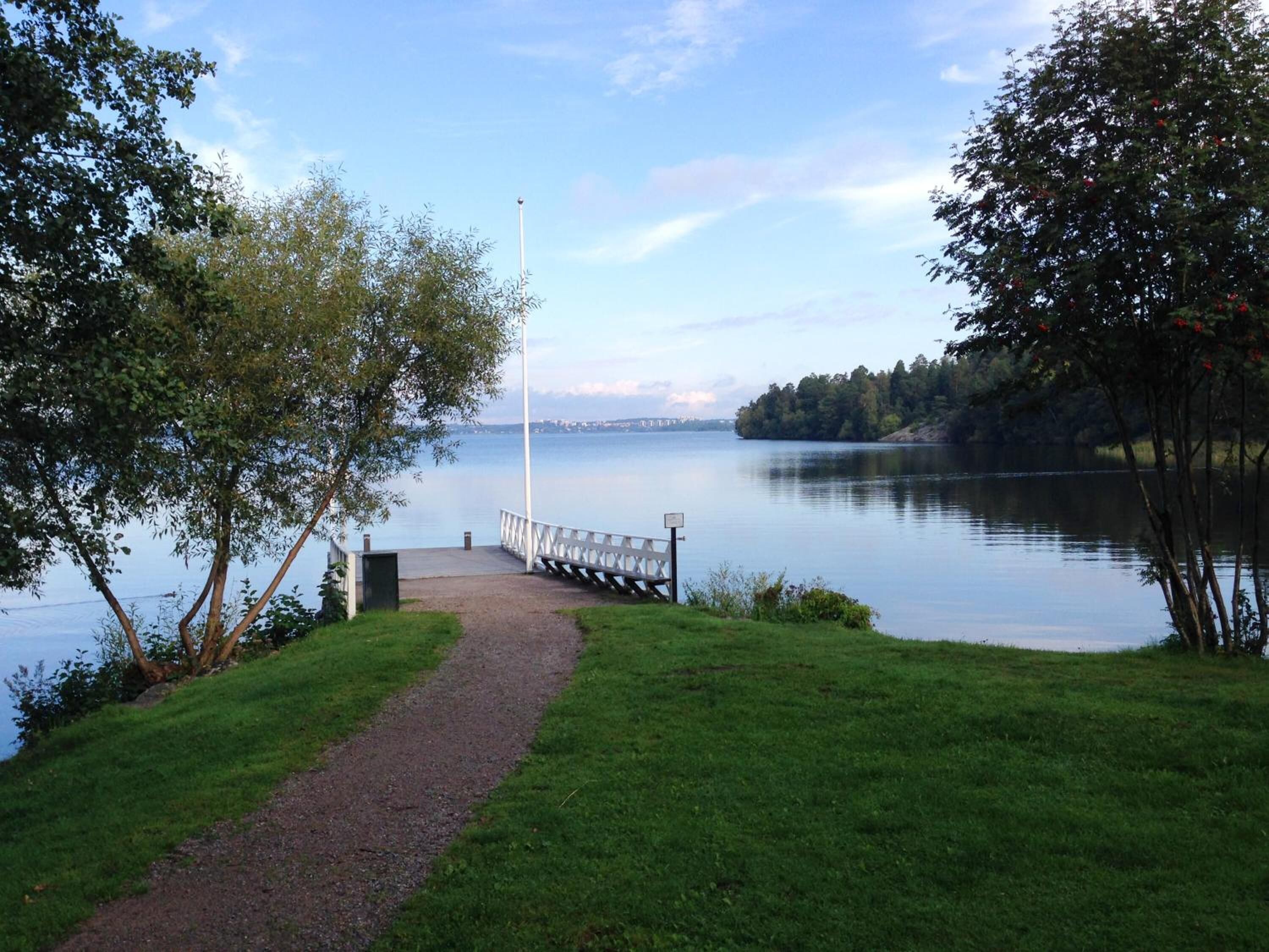 A lakeside path leading to a dock with a flagpole, surrounded by trees and a calm lake.
