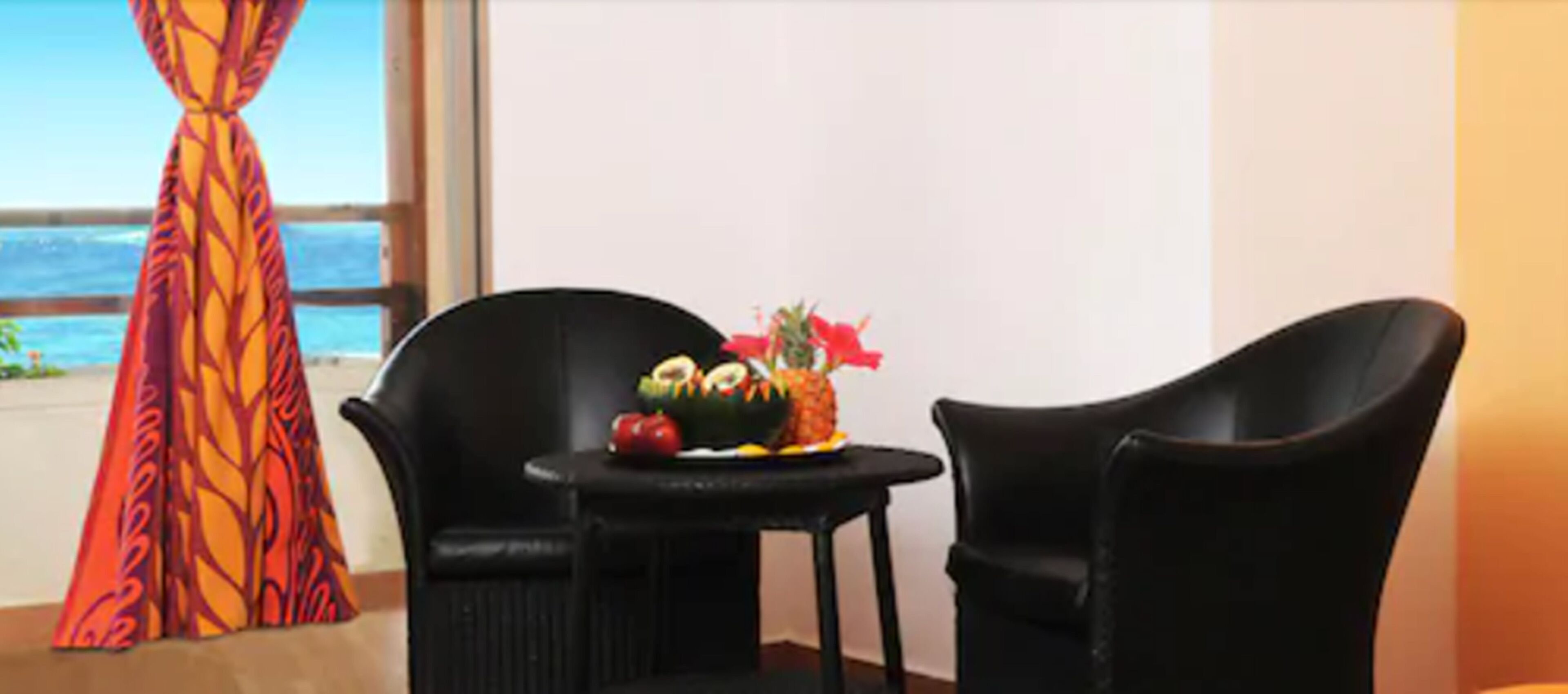 A room with a view of the ocean, a table with fruit and flowers, and two black chairs.
