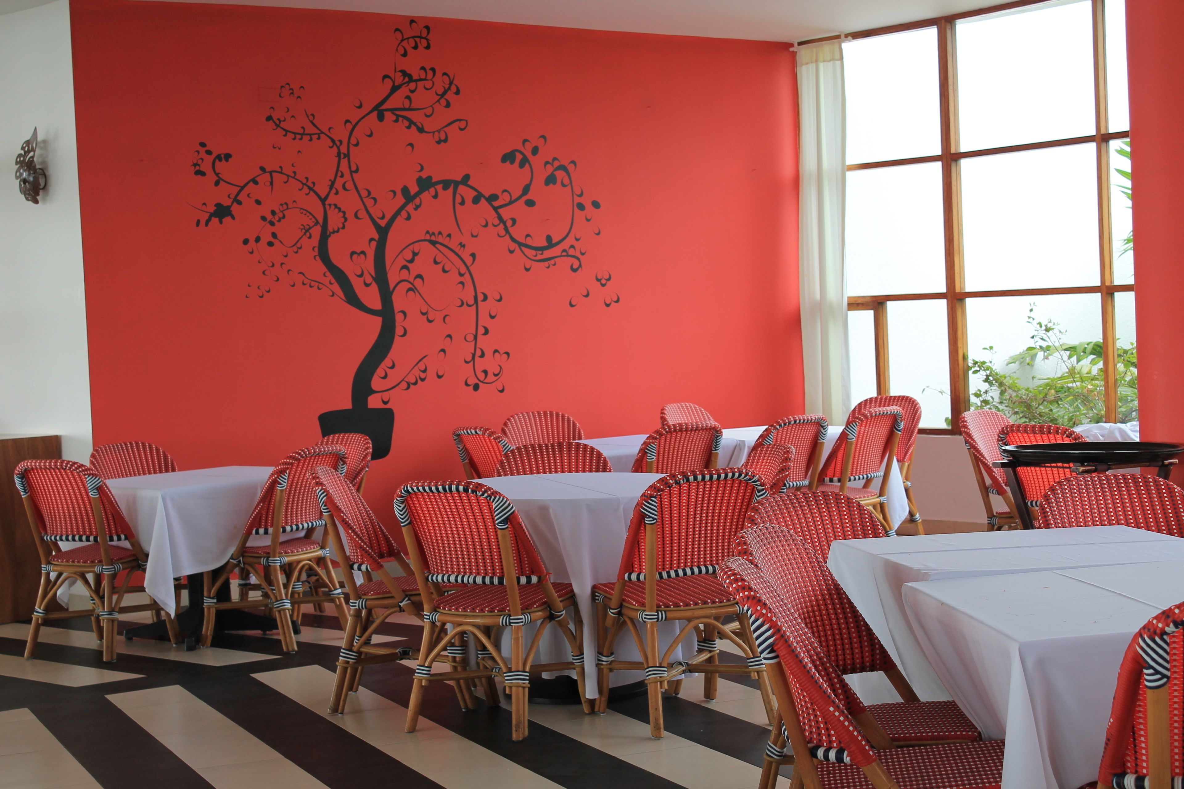 A dining area with red and white checkered chairs, white tablecloths, and a large window with a view of greenery.