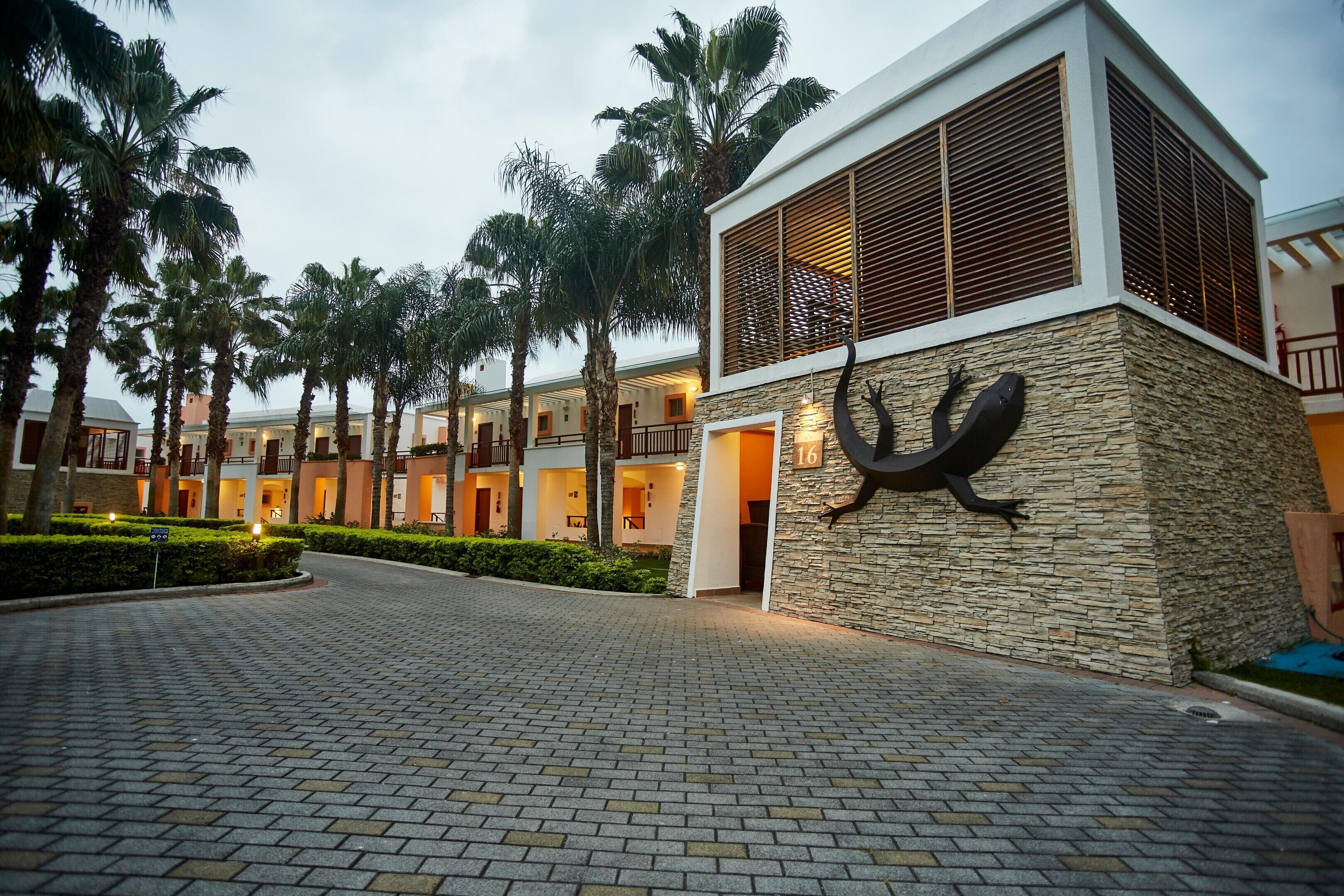 A hotel entrance with a paved driveway, palm trees, and a building with a stone facade and a number sign.