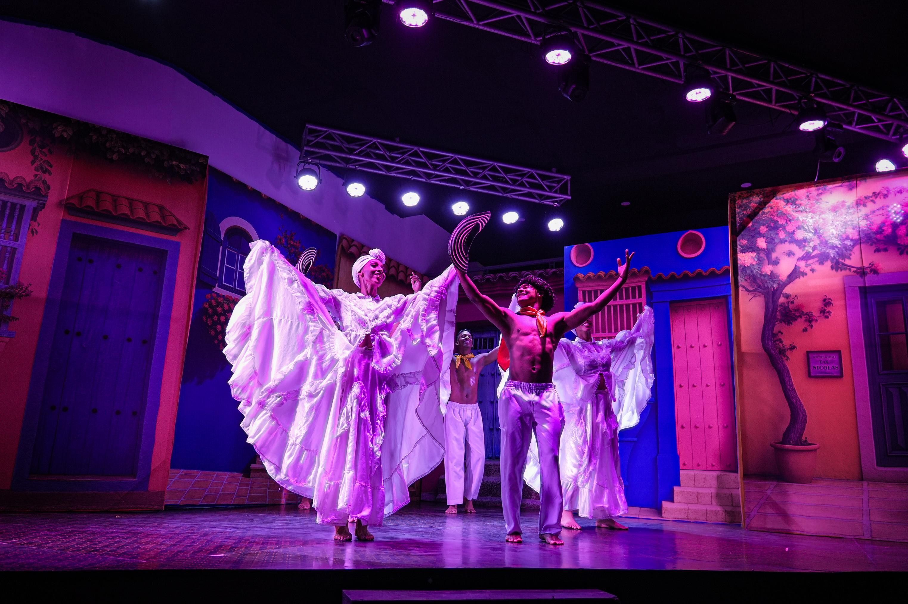 A stage with performers in white costumes, a blue door, and a tree mural.