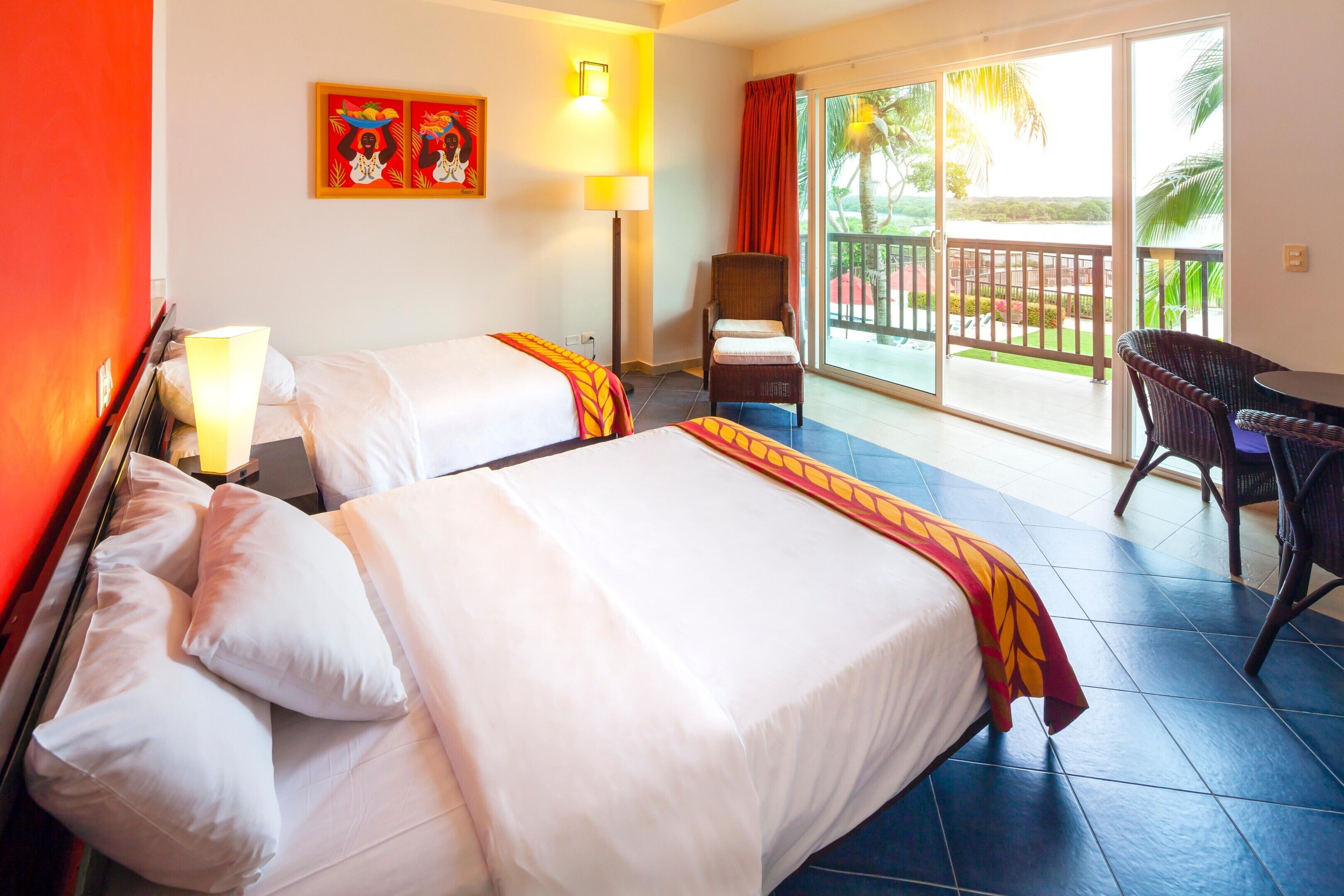 A hotel room with two beds, a chair, a table, a balcony with a view, and a painting on the wall.