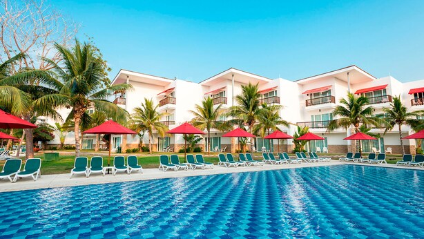 A hotel pool area with multiple lounge chairs and red umbrellas, surrounded by palm trees and a clear blue sky.