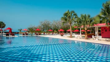 A pool with a checkered pattern, surrounded by lounge chairs and red umbrellas, with palm trees and a clear sky in the background.