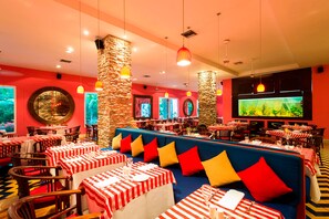Restaurante com mesas listradas em vermelho e branco, assentos acolchoados azuis e pilares de pedra.