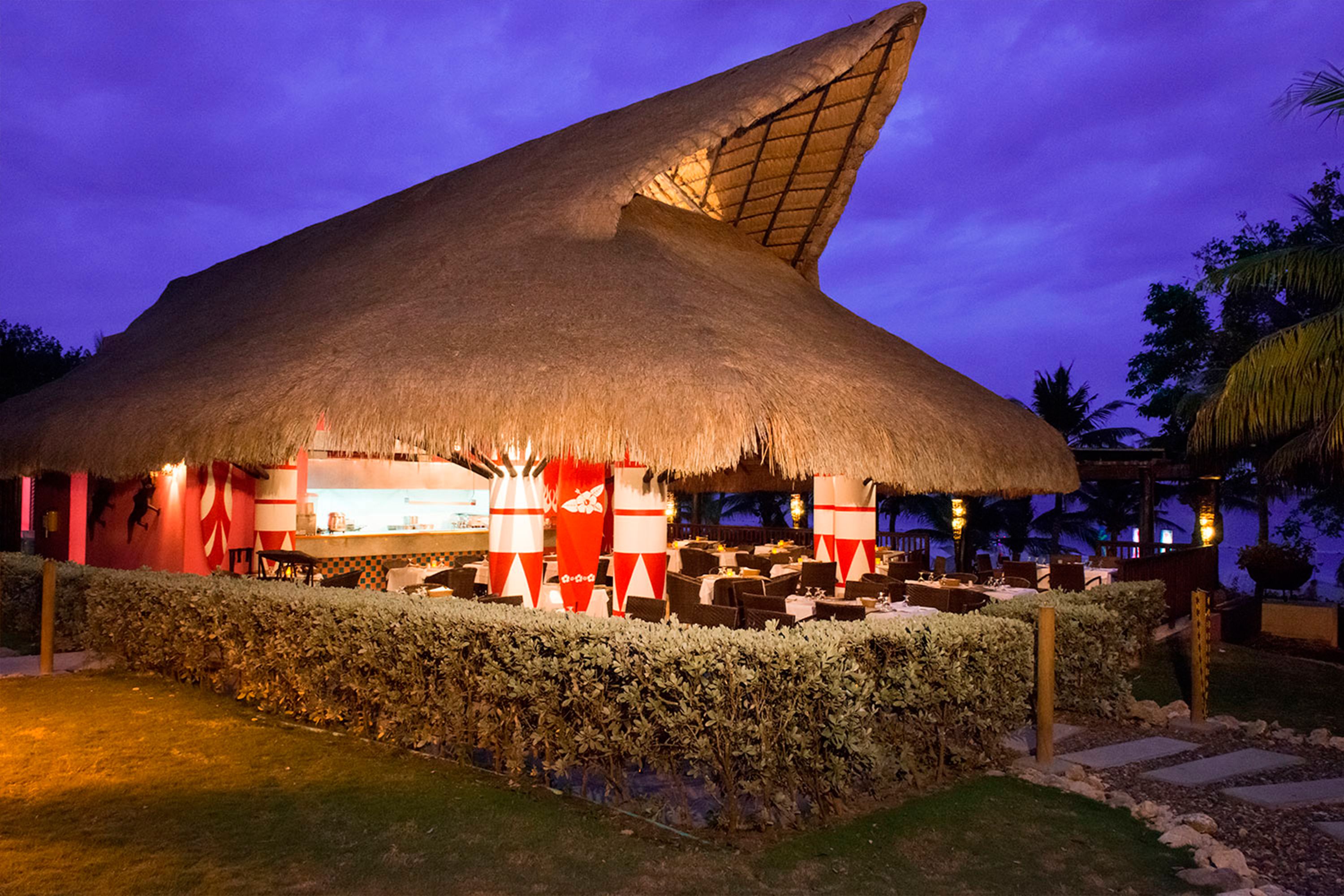A thatched-roof structure with tables and chairs, likely a restaurant or lounge area, surrounded by hedges and palm trees.