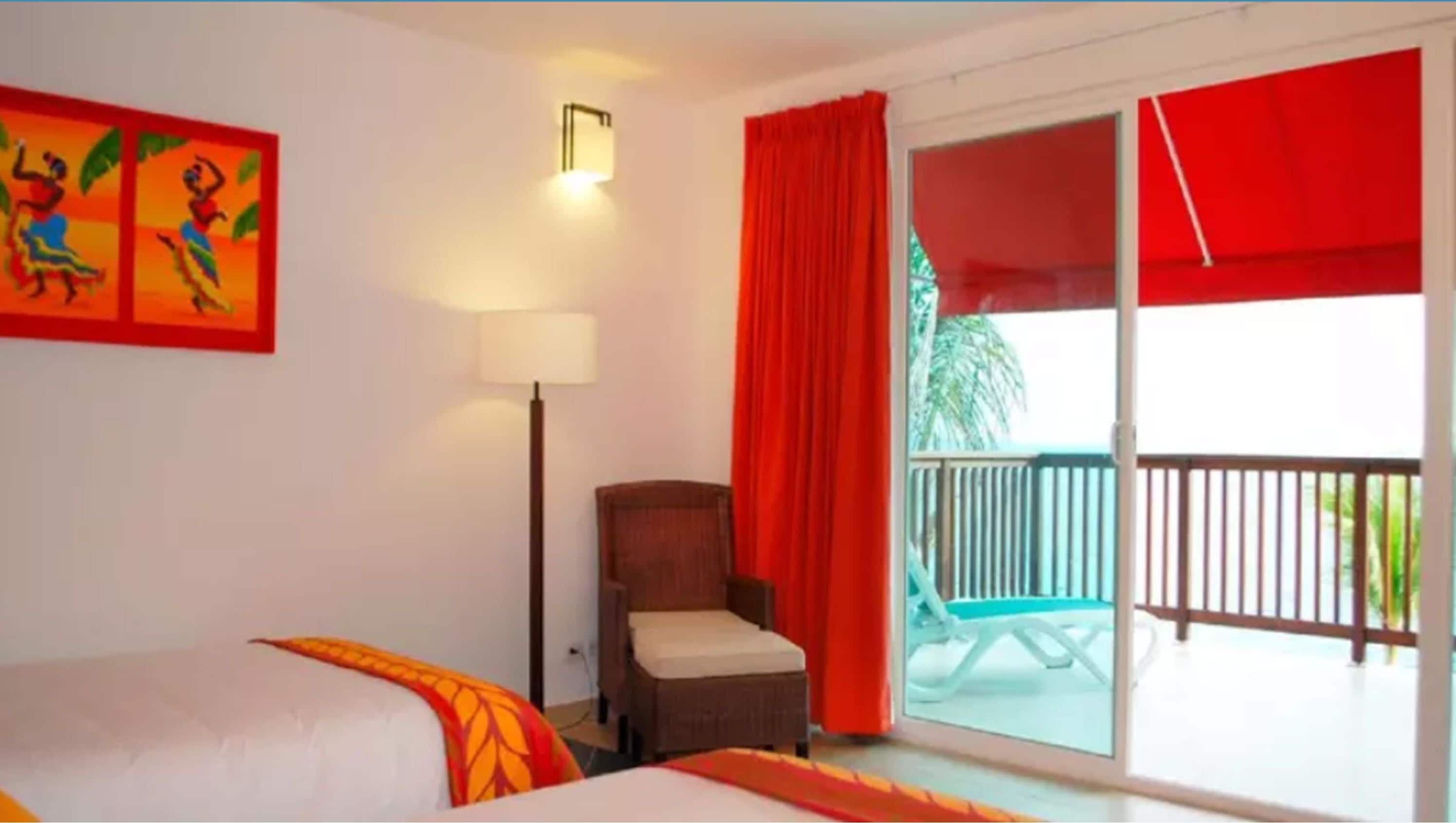 A hotel room with a bed, a chair, a balcony with red curtains, and a painting on the wall.