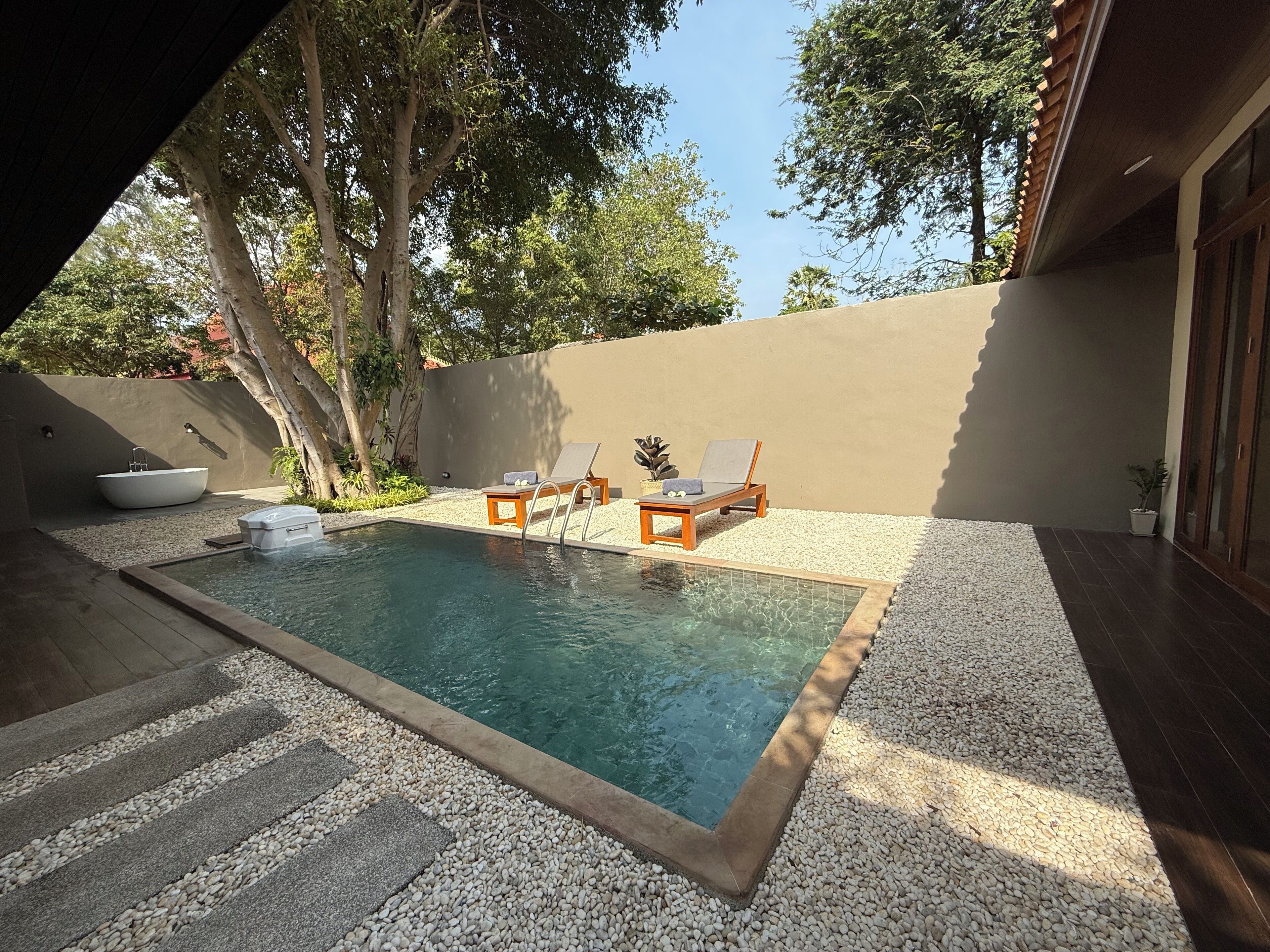 A rectangular swimming pool with two lounge chairs and a small table, surrounded by pebbles and a low wall.