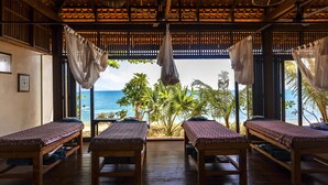 A room with wooden floors, four beds with patterned bedspreads, a ceiling fan, and a view of the ocean through a window with curtains. - Saree Samui Natures Finest Resort (Koh Samui)