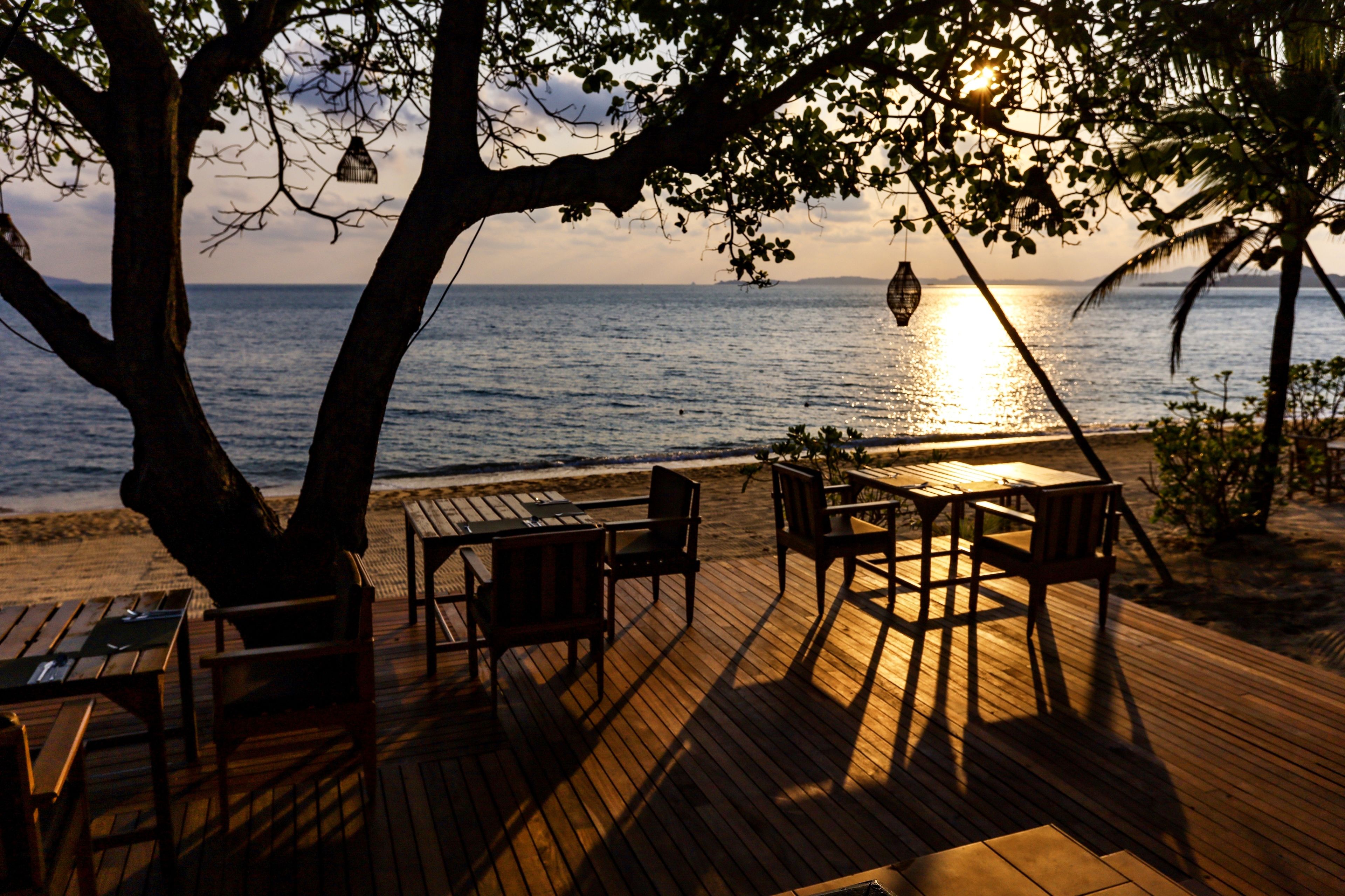 Una terraza de madera con mesas y sillas, con vista a una playa donde se ve el océano y la puesta de sol.