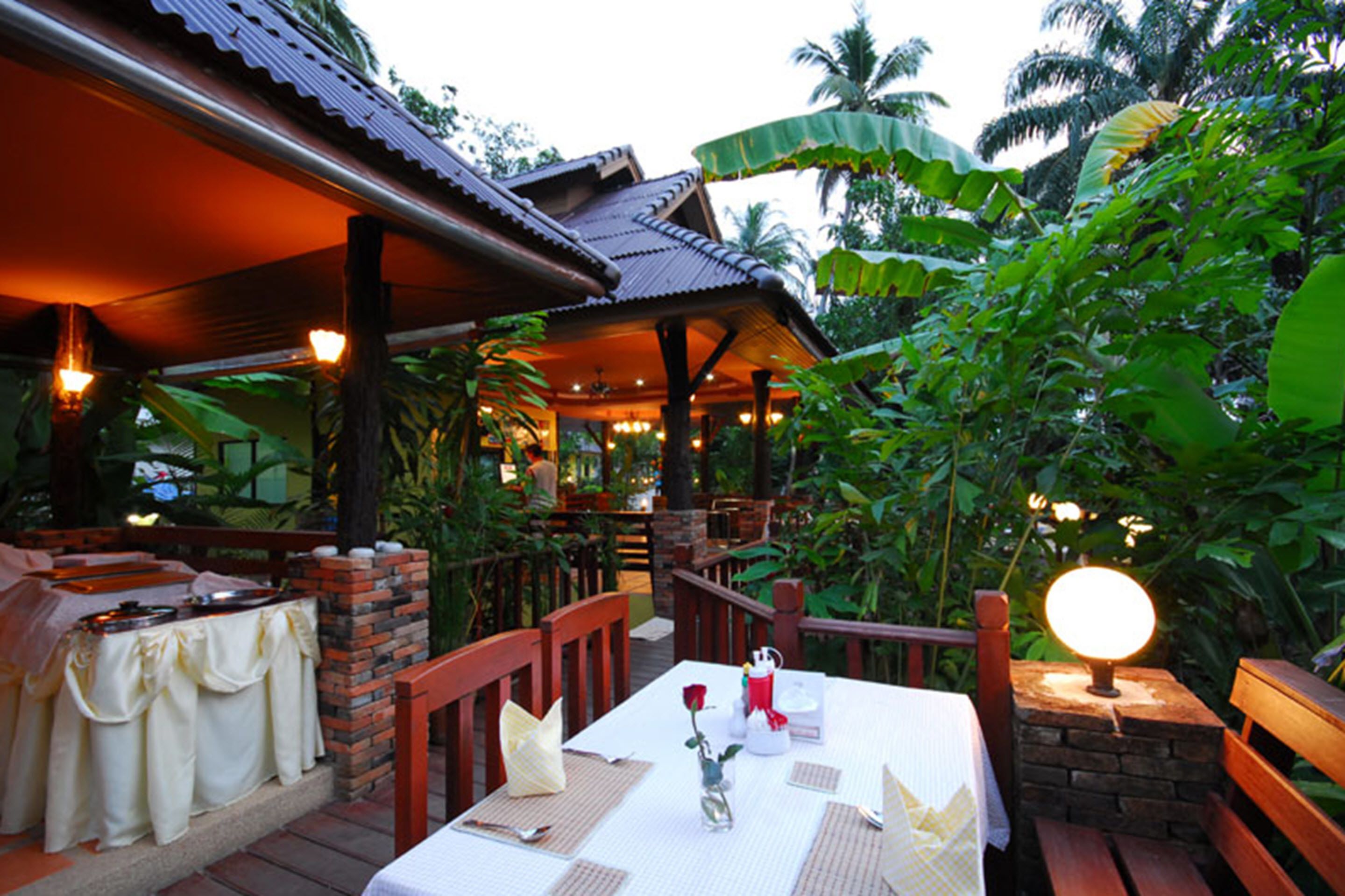 An outdoor dining area with tables set for two, surrounded by lush greenery and a covered seating area.