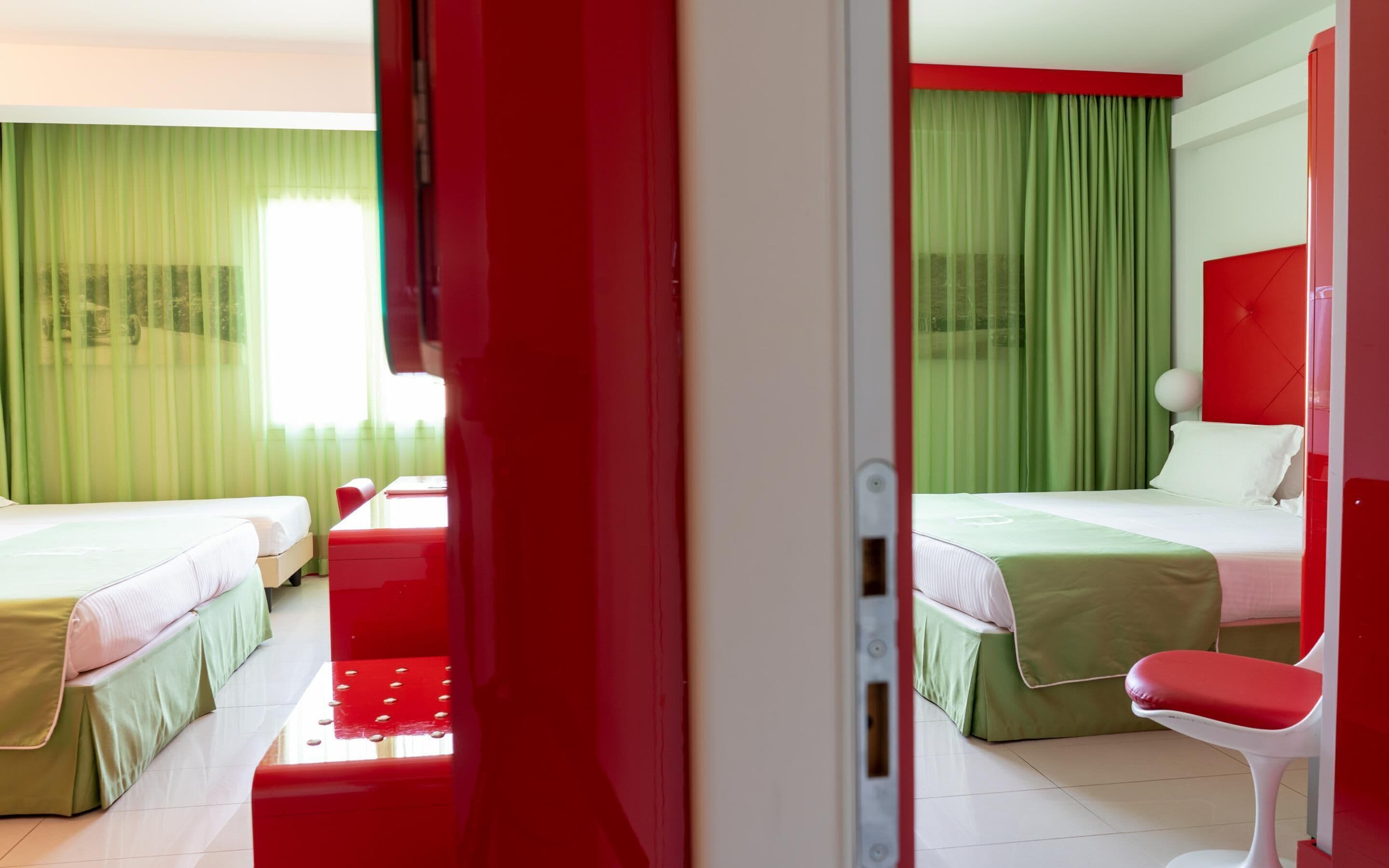 A hotel room with two beds, green curtains, a red accent wall, and a white chair.