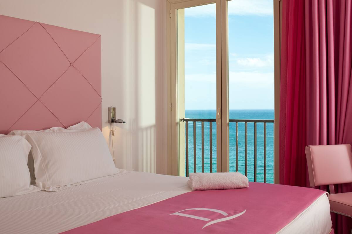 A hotel room with a bed, a view of the ocean, and a balcony with red curtains.