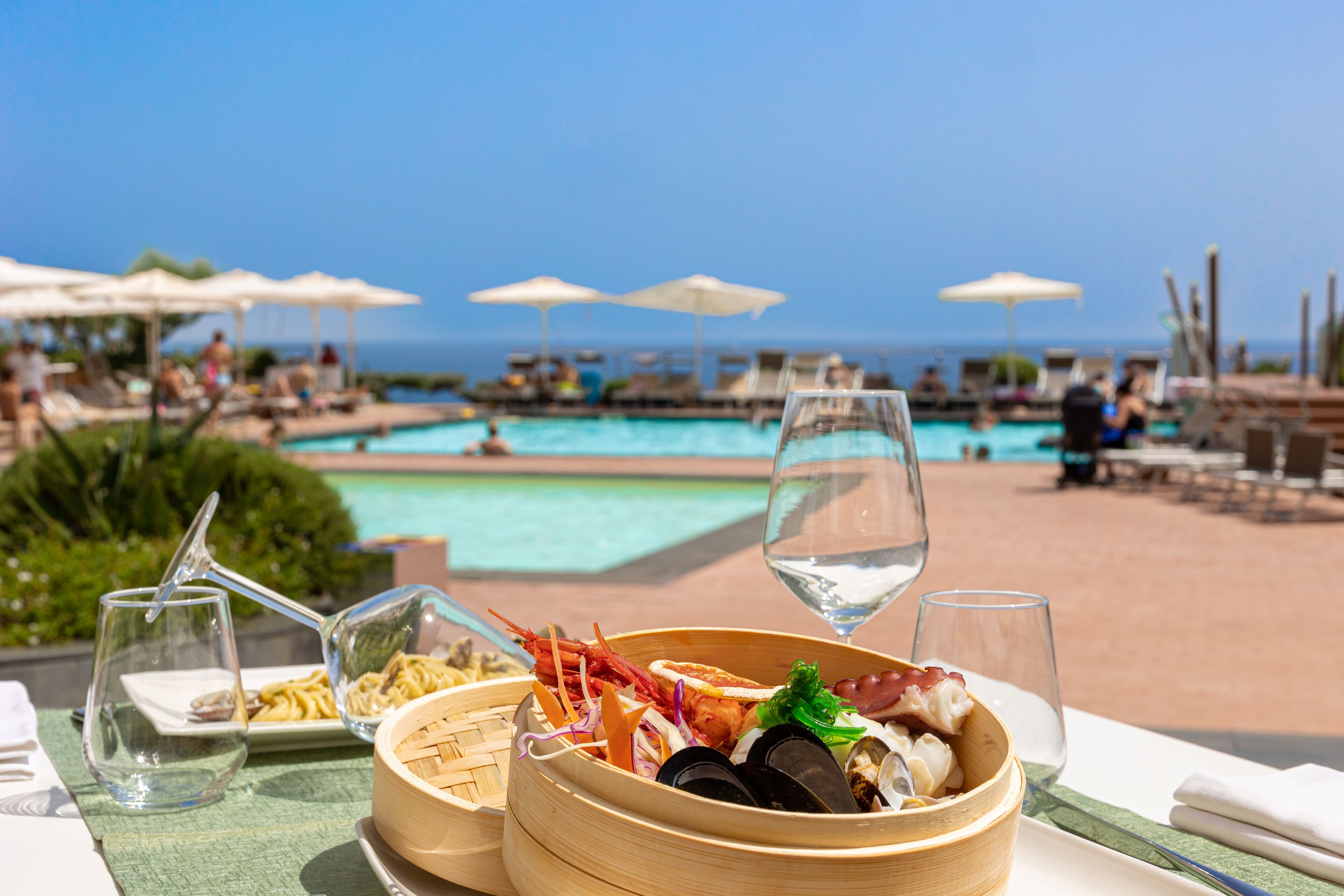 Un tavolo da pranzo con piatti di pesce, un bicchiere d'acqua e vista su una piscina e sdraie.