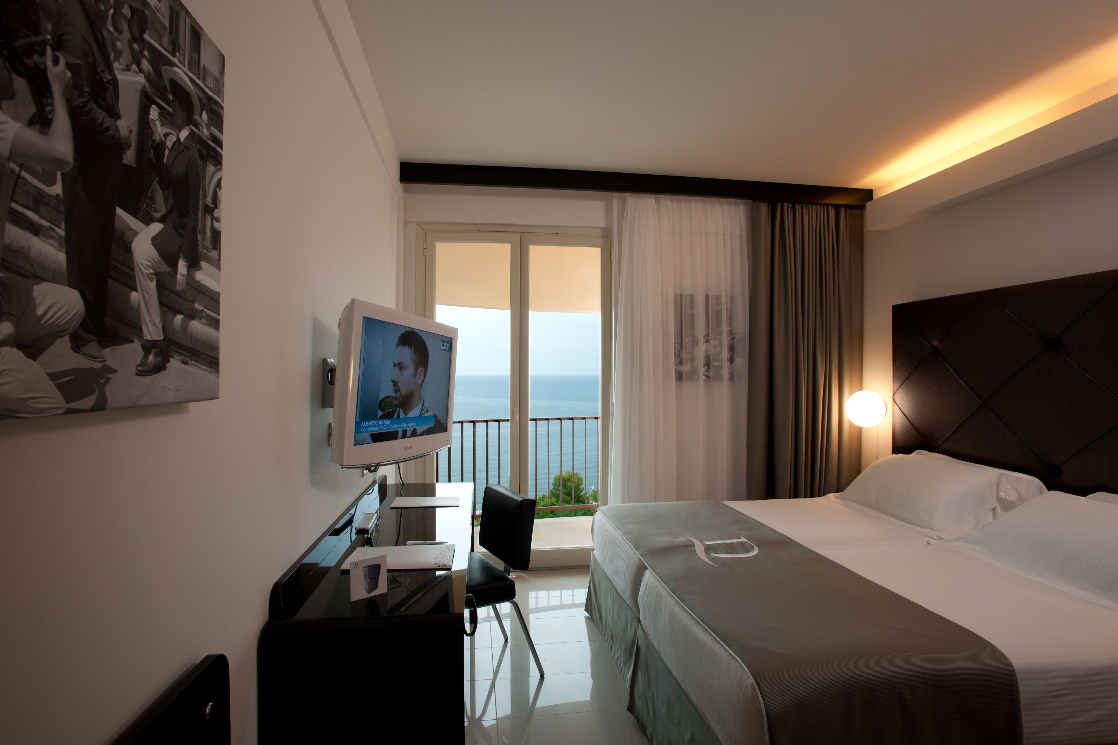 A hotel room with a bed, a desk with a computer, a television, and a balcony with a view of the sea.