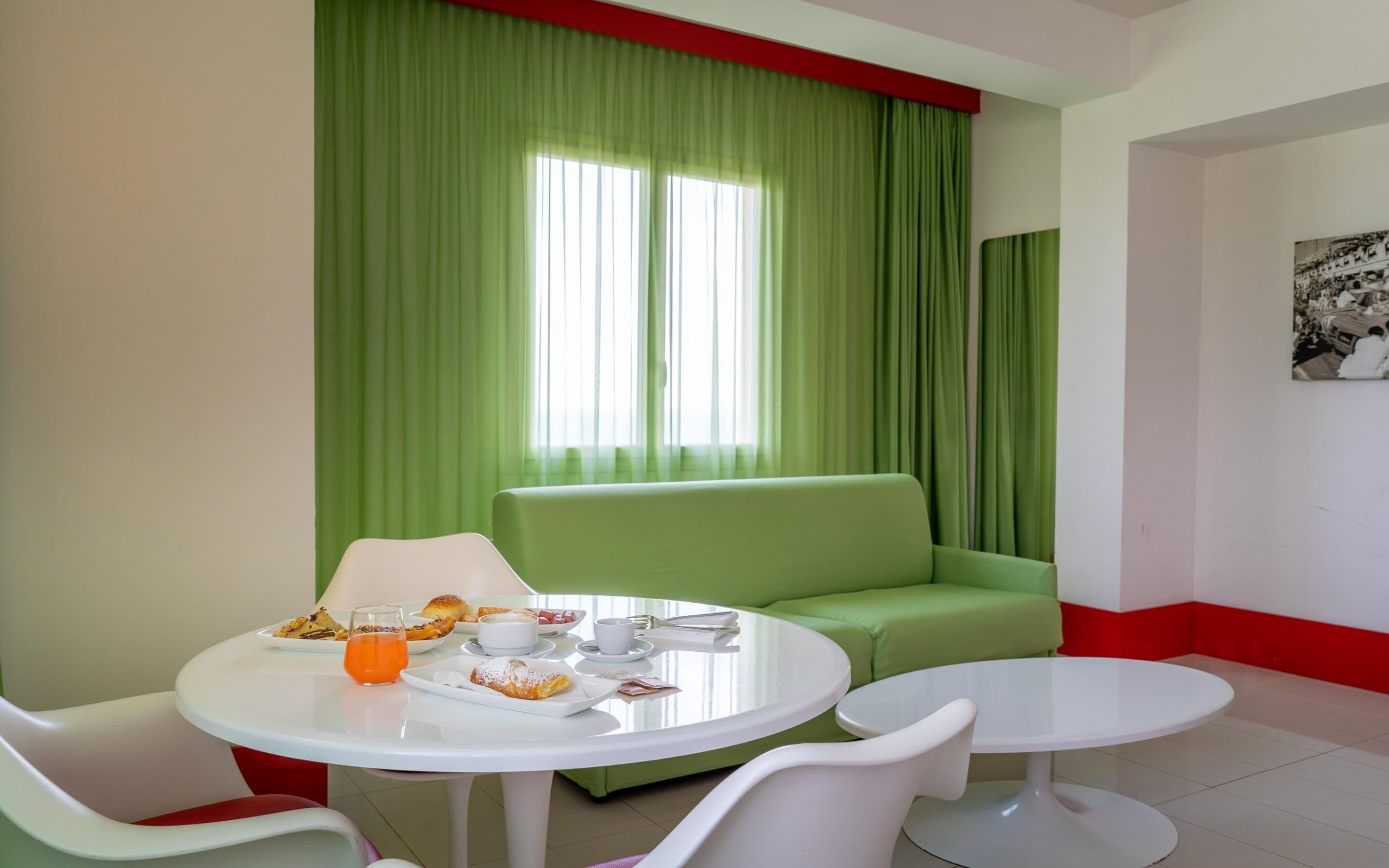 A modern room with a green sofa, two white round tables, and a breakfast setup including pastries and drinks.