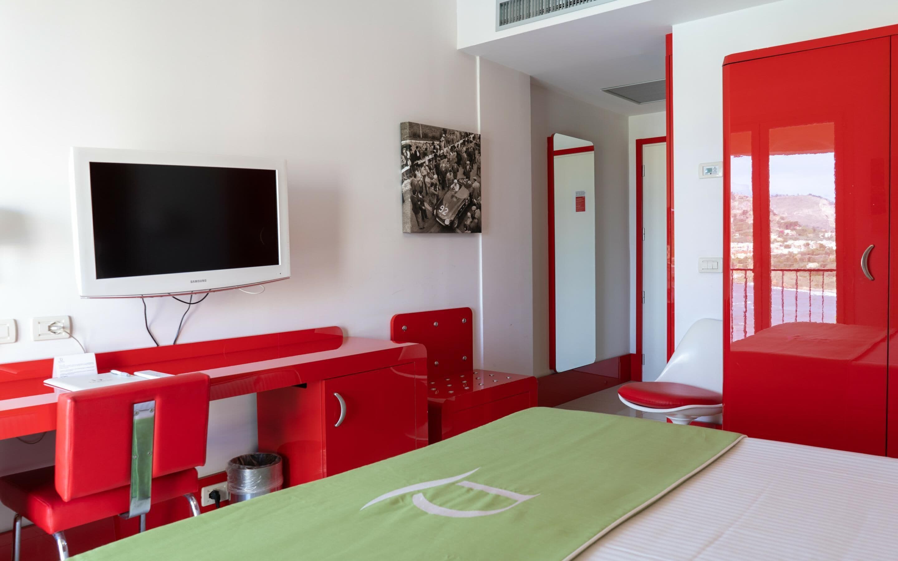 A hotel room with a red desk, a television, a bed with a green bedspread, and a view of the beach.