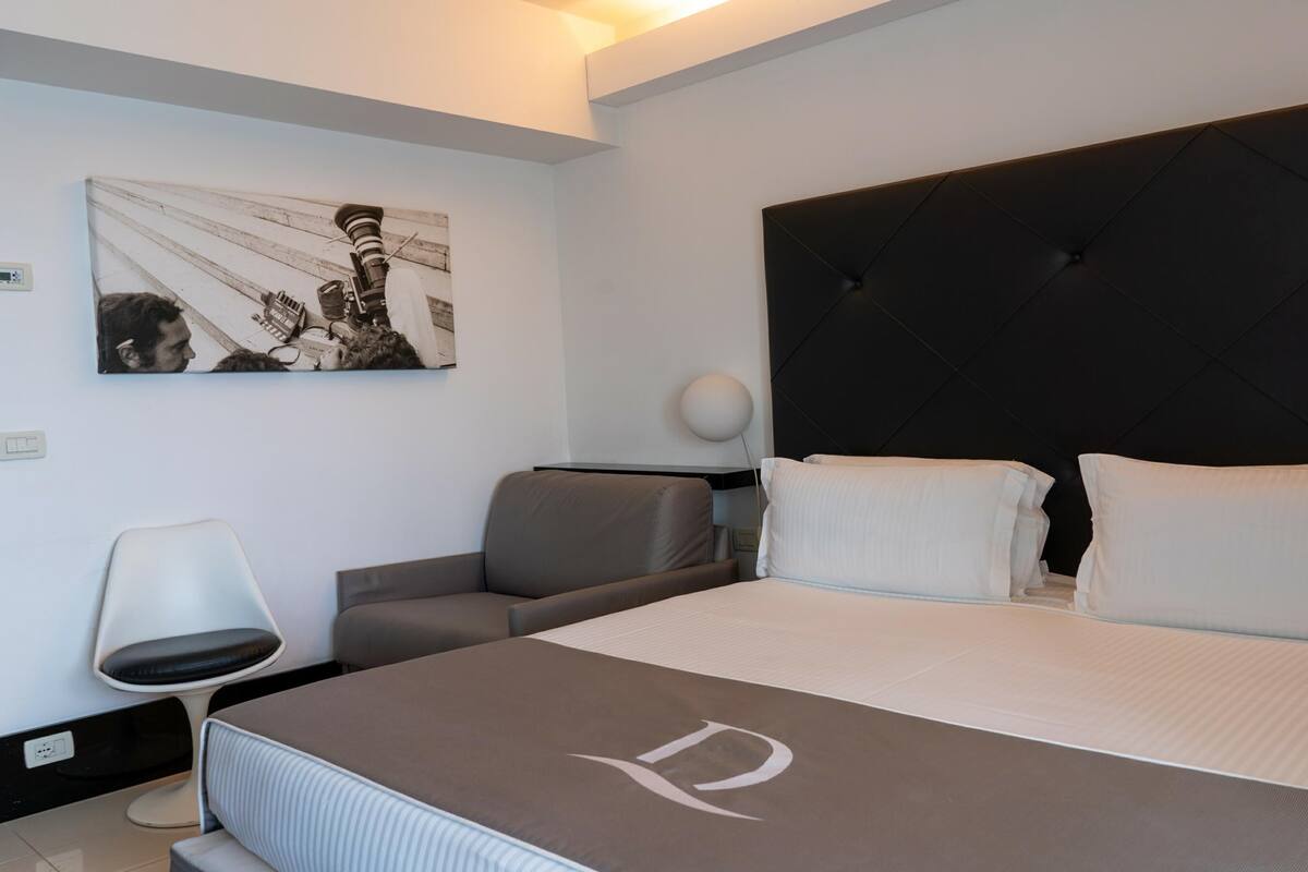 A modern hotel room with a large bed, a headboard, a desk, a chair, and a wall-mounted artwork.