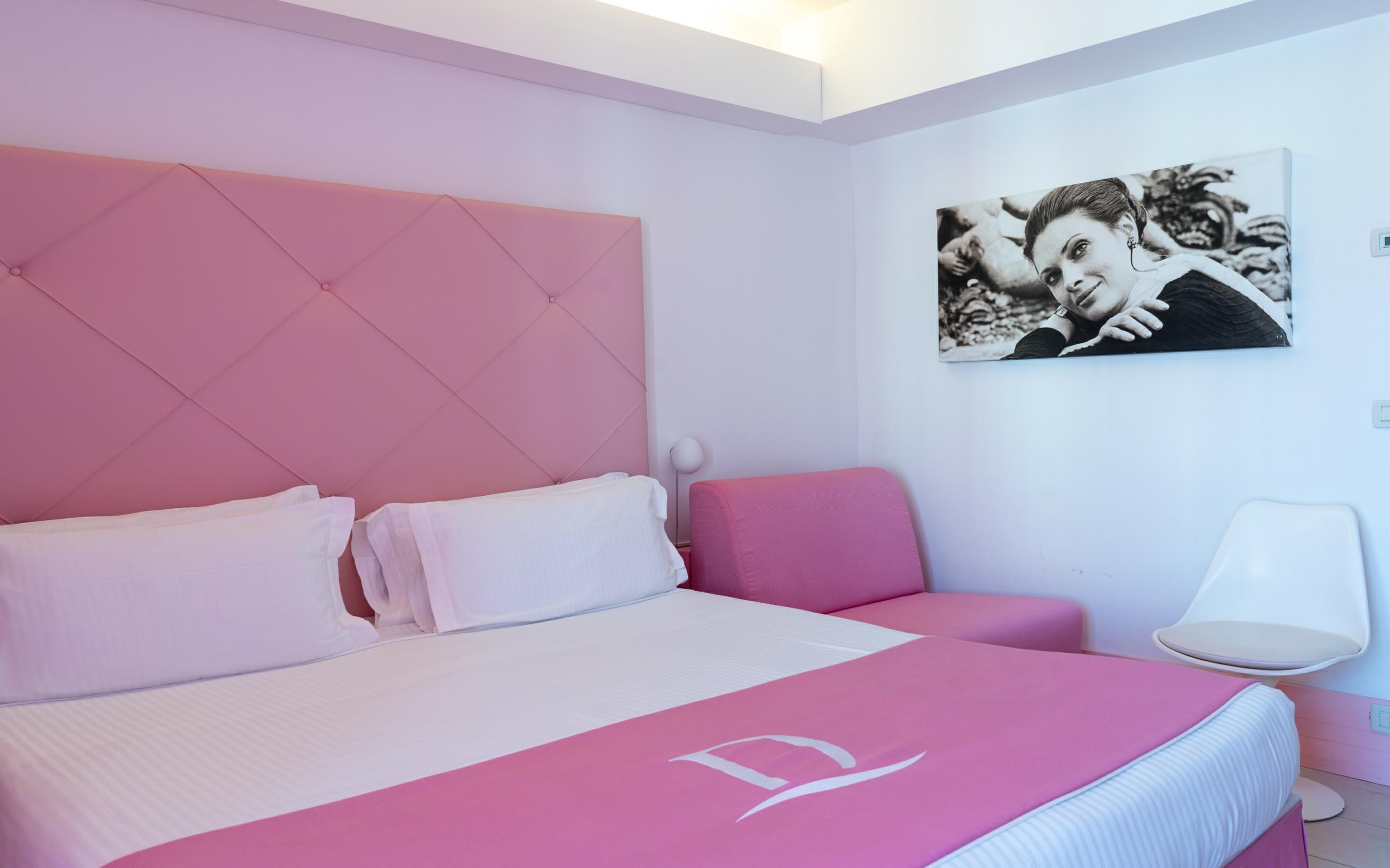 A modern bedroom with a pink and white color scheme, featuring a bed with a tufted headboard, a bedside table, a chair, and a wall-mounted picture.