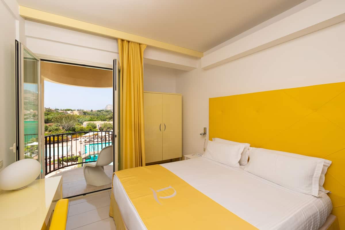 A hotel room with a bed, a desk, a chair, and a view of a pool and greenery.
