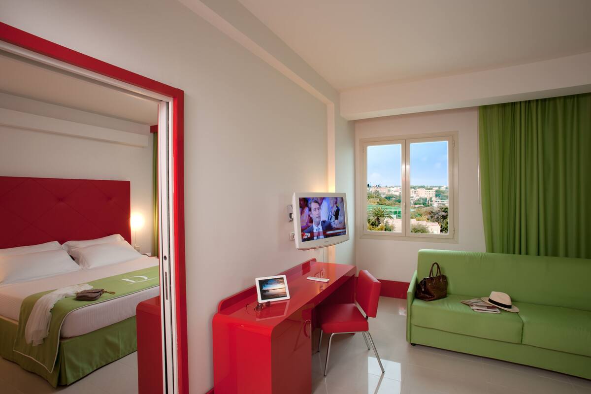 A hotel room with a bed, a desk with a computer, a green sofa, and a TV.