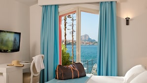 A hotel room with a bed, a desk, a TV, and a view of the sea and a mountain. - Domina Zagarella Sicily (Santa Flavia)