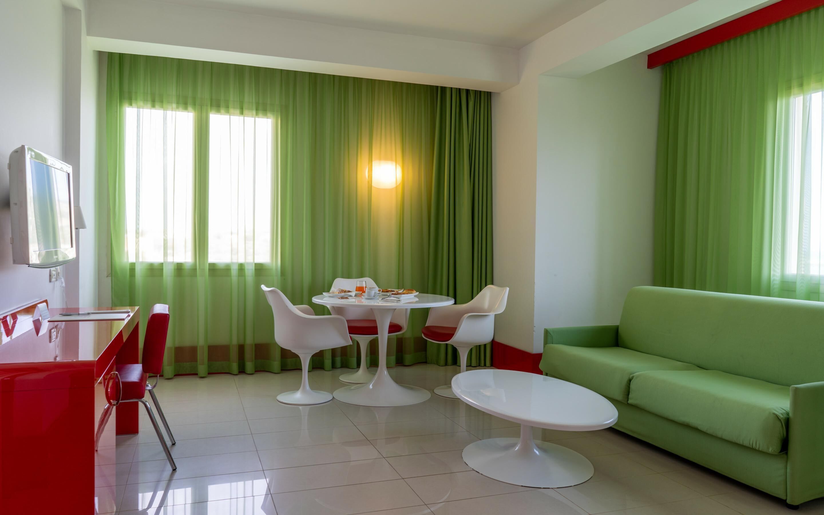 A modern hotel room with a dining area featuring a round table and four chairs, a green sofa, and a TV mounted on the wall.