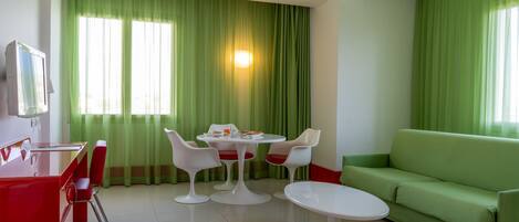 A modern hotel room with a dining area featuring a round table and four chairs, a green sofa, and a TV mounted on the wall.
