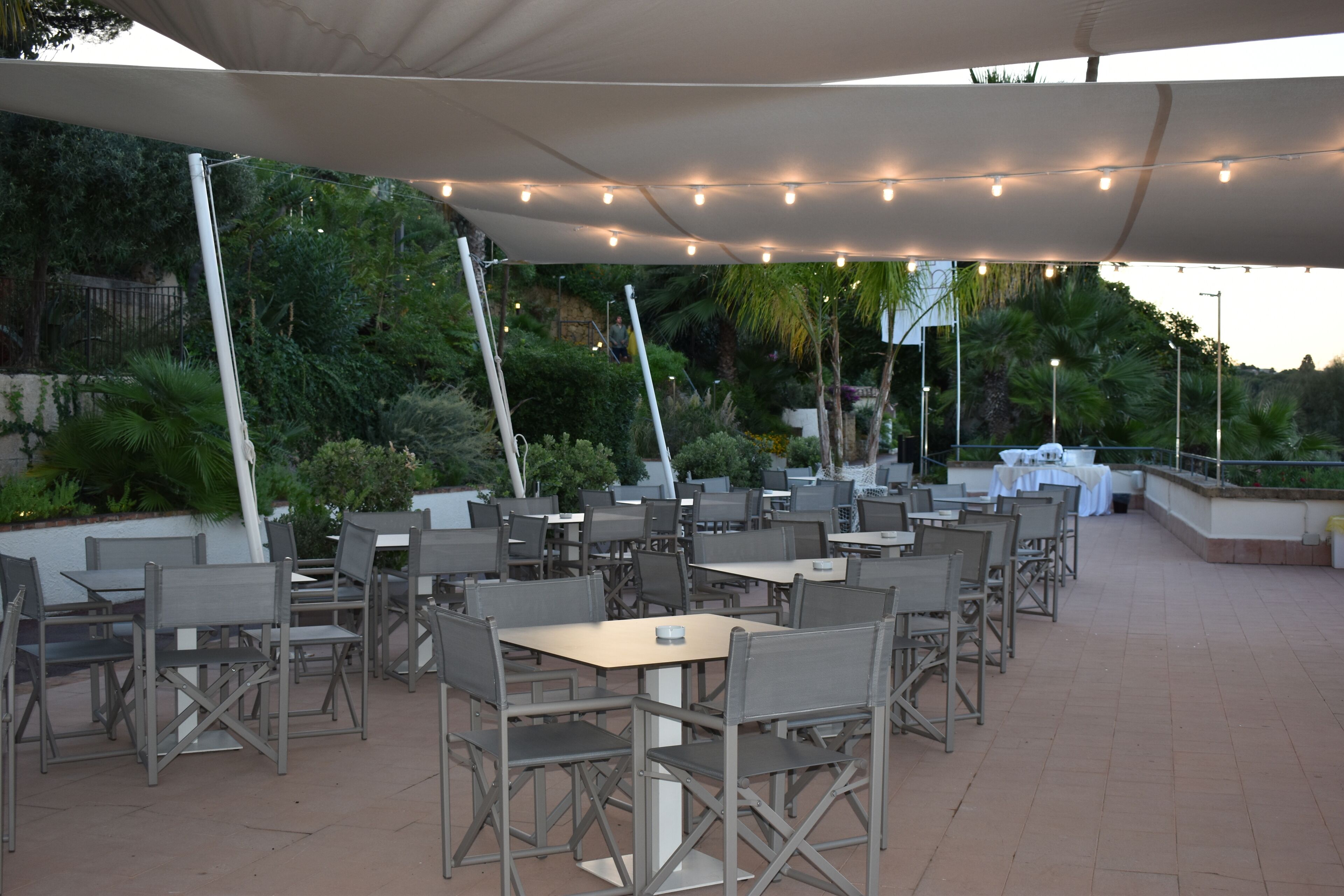 An outdoor patio with multiple tables and chairs, surrounded by greenery and a white canopy.