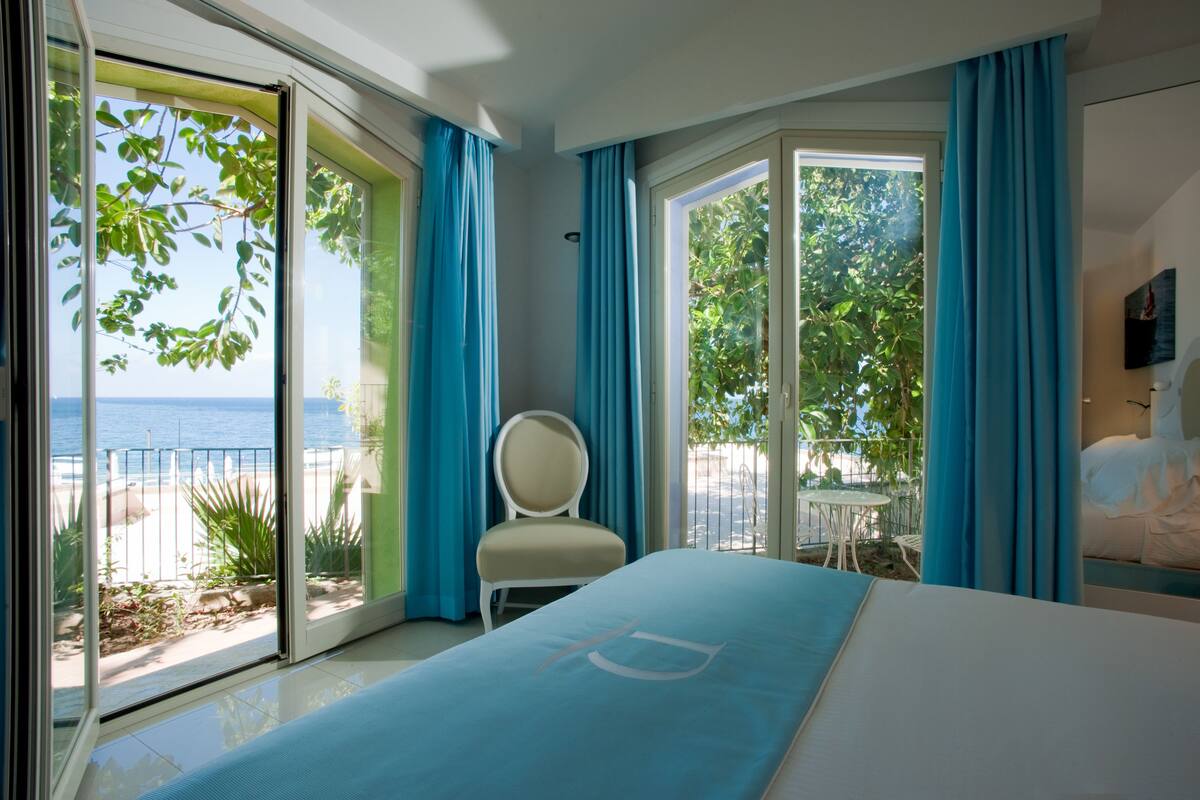A hotel room with a bed, a chair, and a view of the sea through large windows.