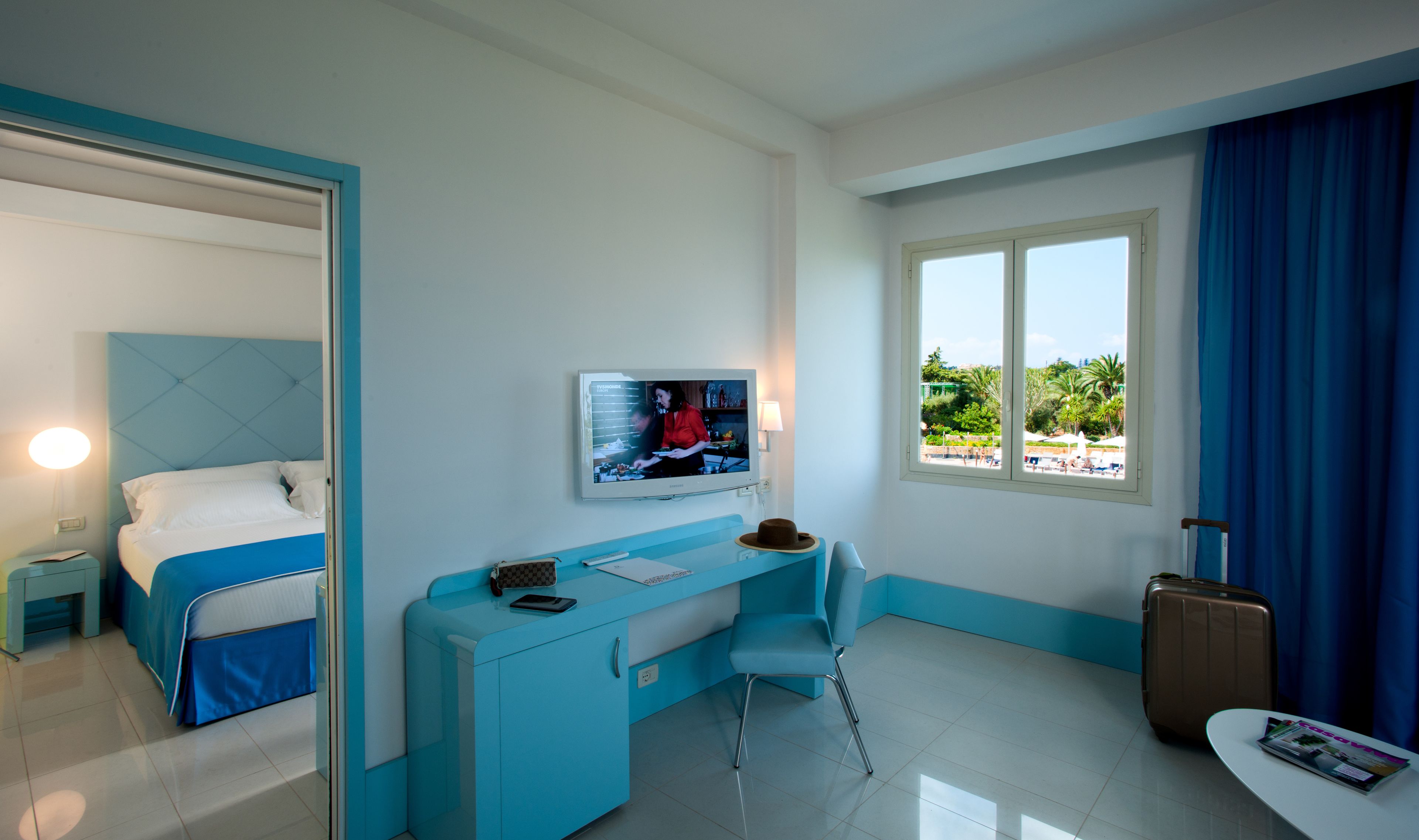 Une chambre d’hôtel avec un lit, un bureau, une chaise, une télévision, une fenêtre donnant sur une piscine et une valise.