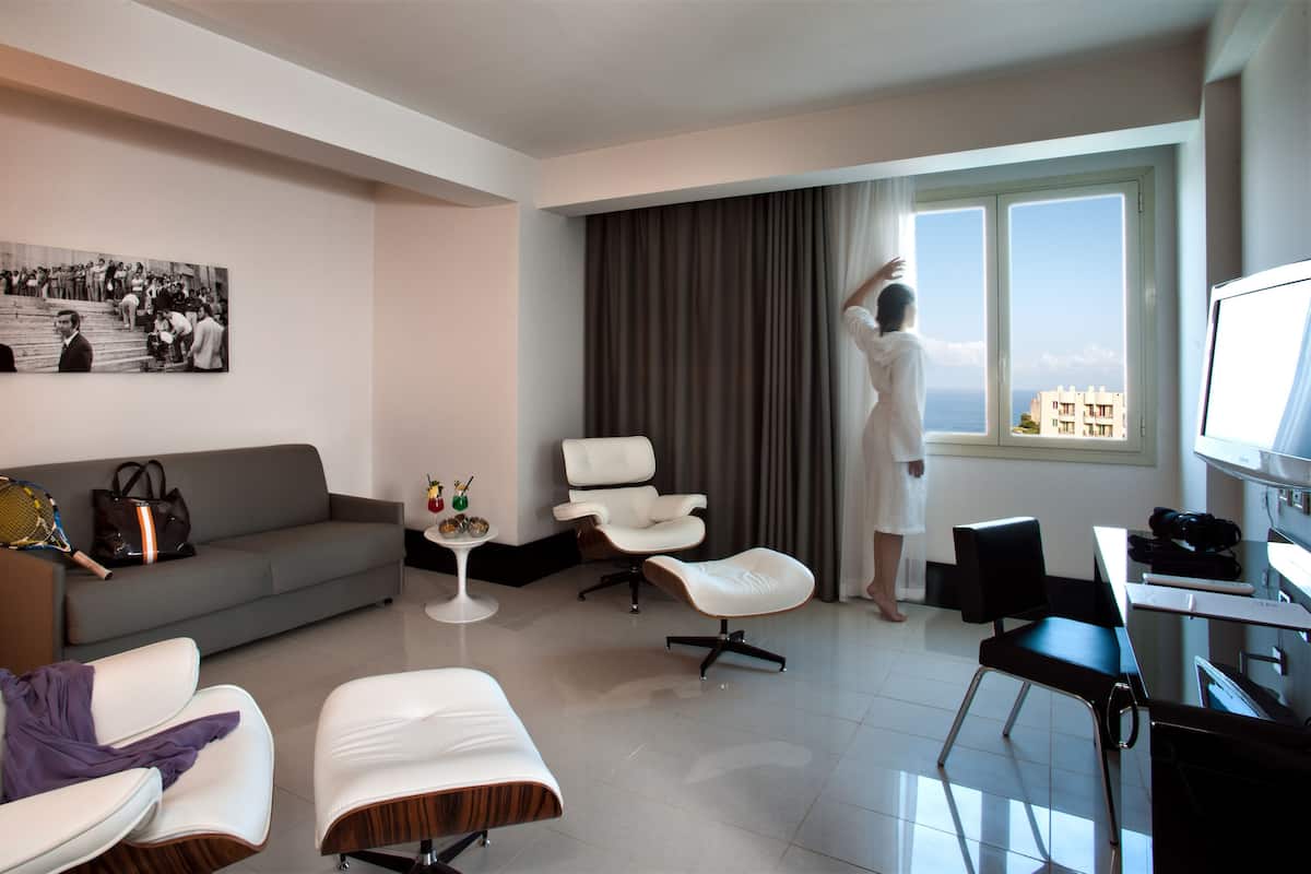 A modern hotel room with a grey sofa, two white armchairs, a small side table with drinks, a large window with curtains, and a TV on a desk.