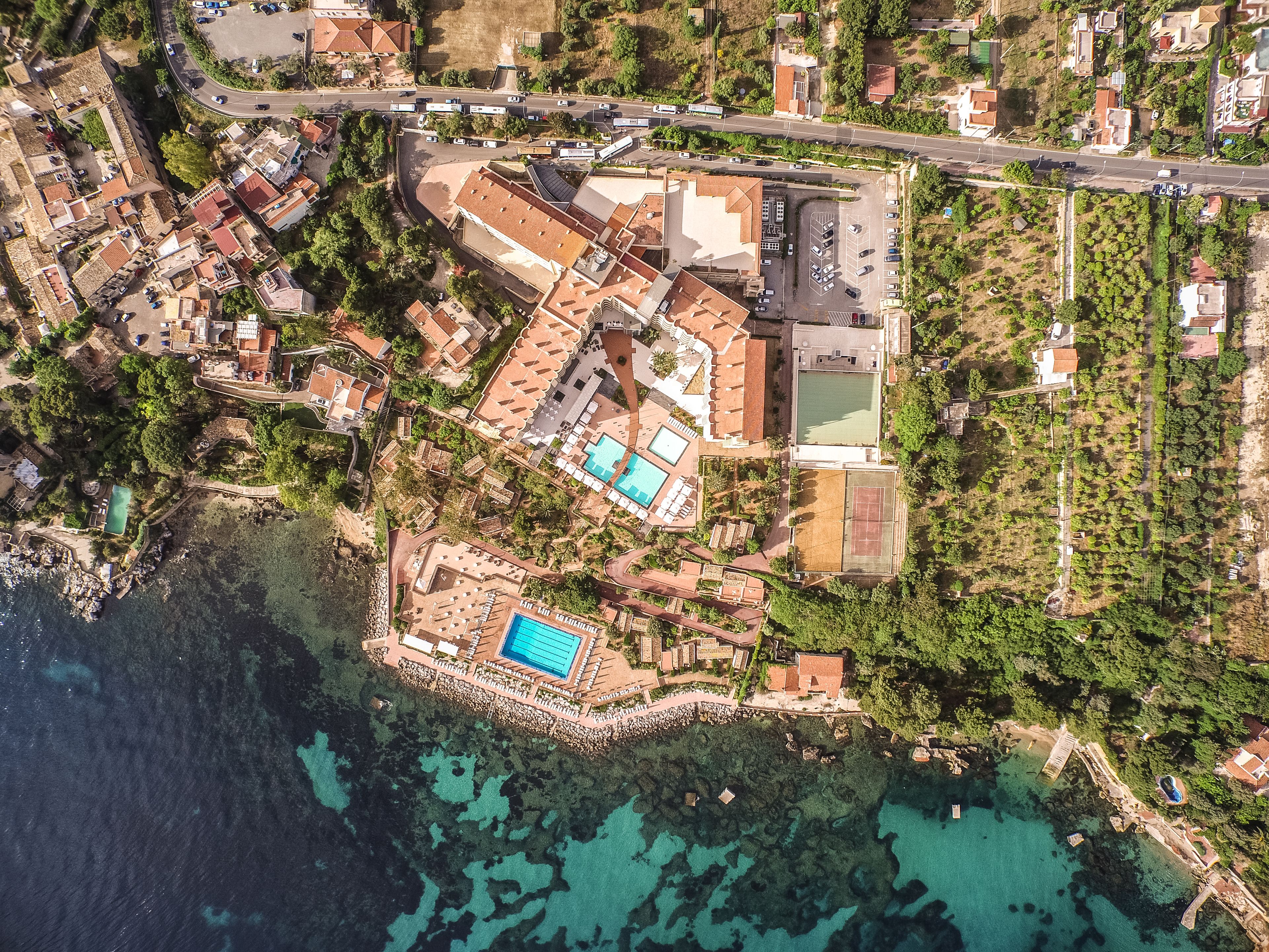 Ein Luftbild eines Küstenresorts mit mehreren Gebäuden, einem großen Schwimmbecken und einem Tennisplatz.