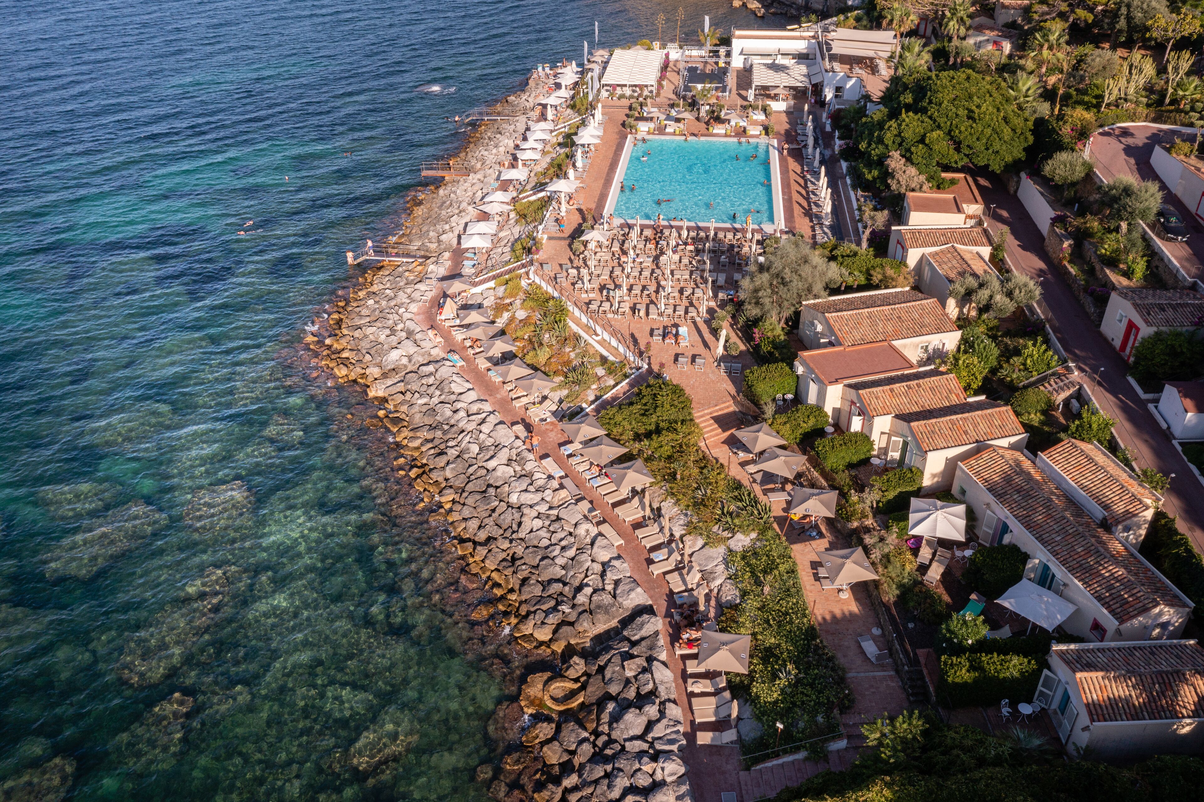 Luftaufnahme eines Küstenresorts mit Schwimmbad, Strandmöbeln und Villen.