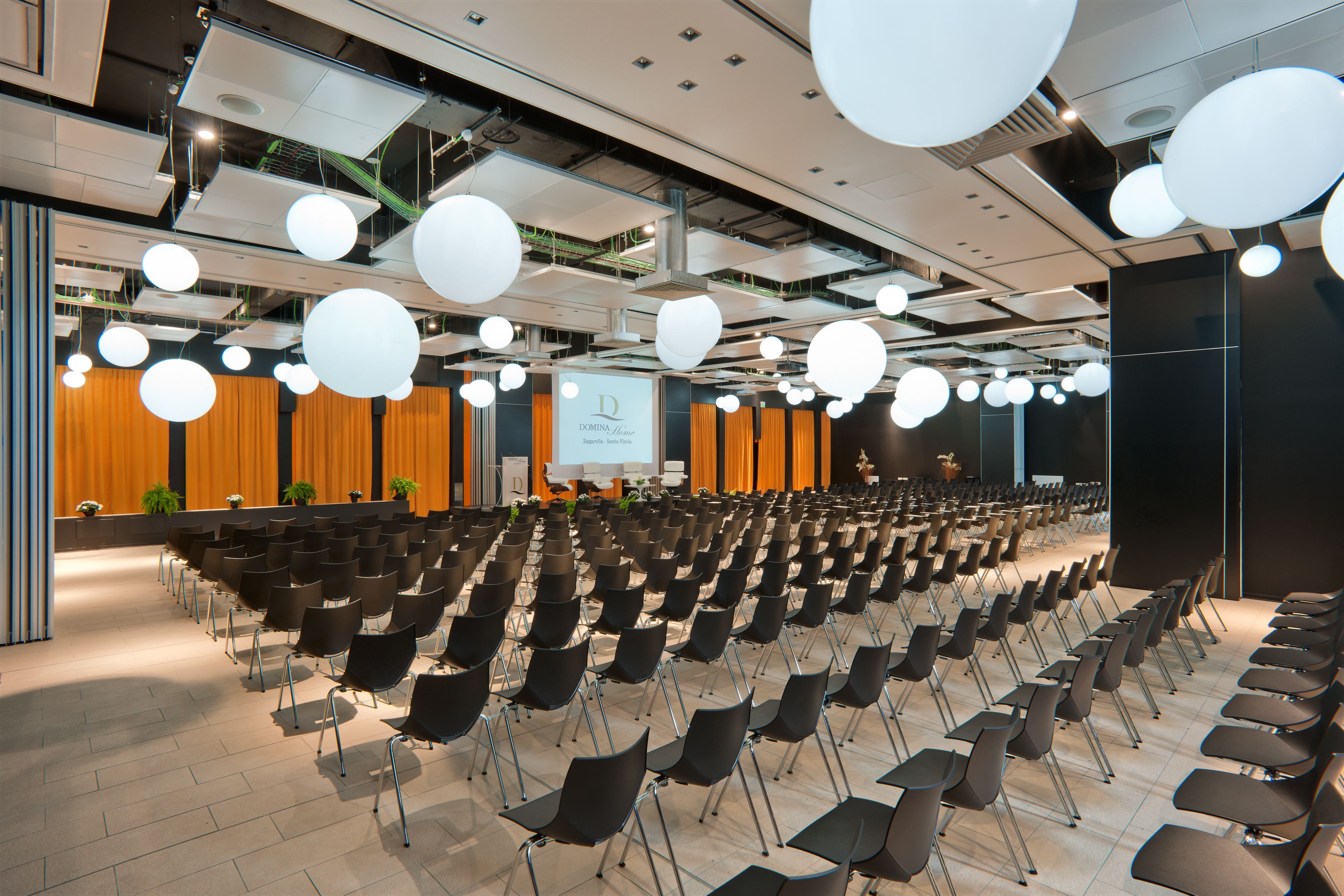 Una sala conferenze con file di sedie, un palco con uno schermo e lampade moderne.