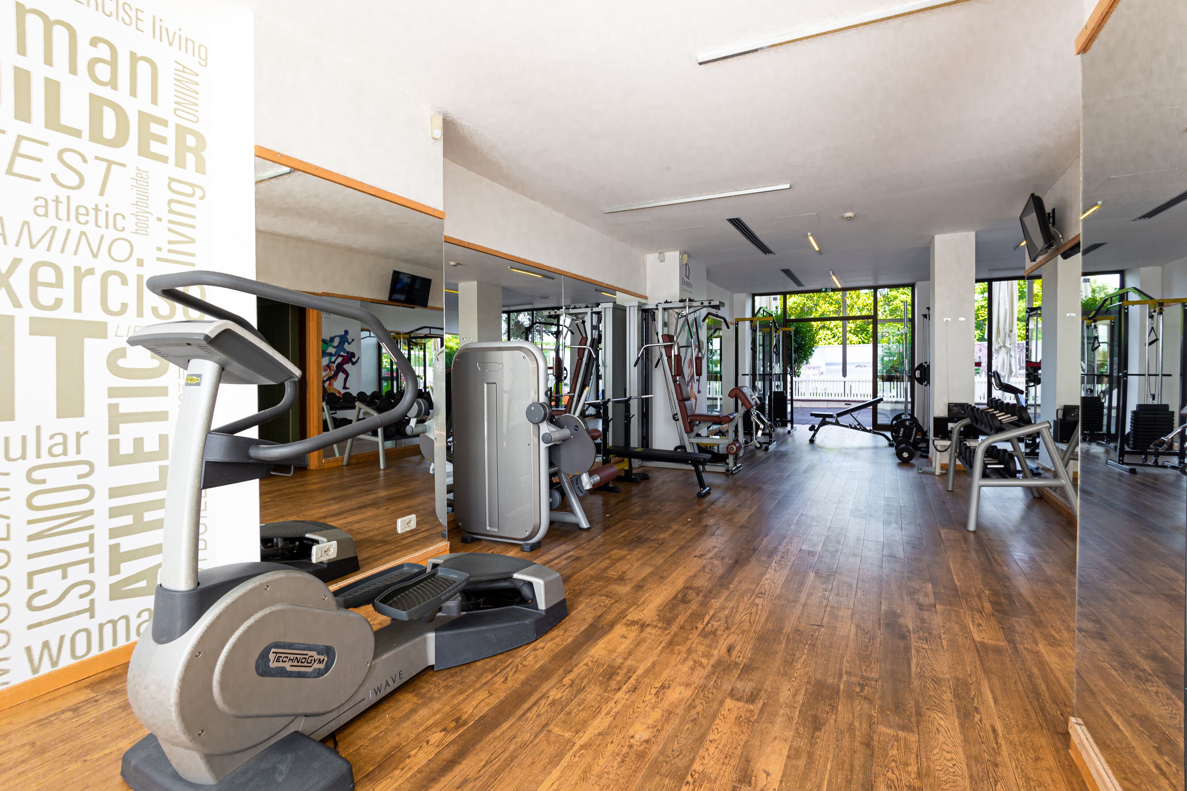 Una palestra con diverse macchine per esercizi, pavimento in legno e ampie finestre che lasciano entrare la luce naturale.