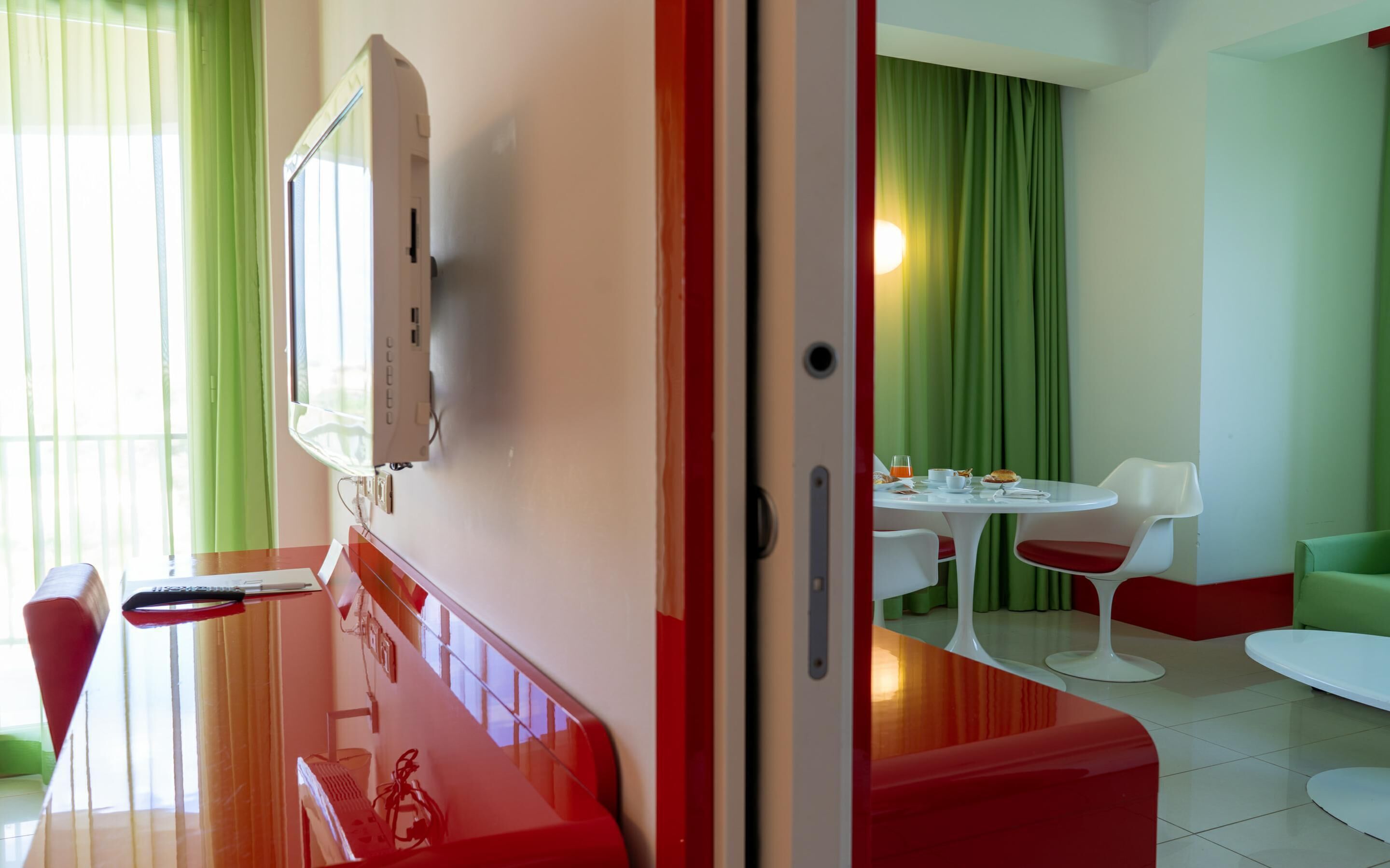 Ett modernt hotellrum med en platt-TV som är monterad på väggen, en matplats med ett bord och stolar samt en grön gardin i bakgrunden.