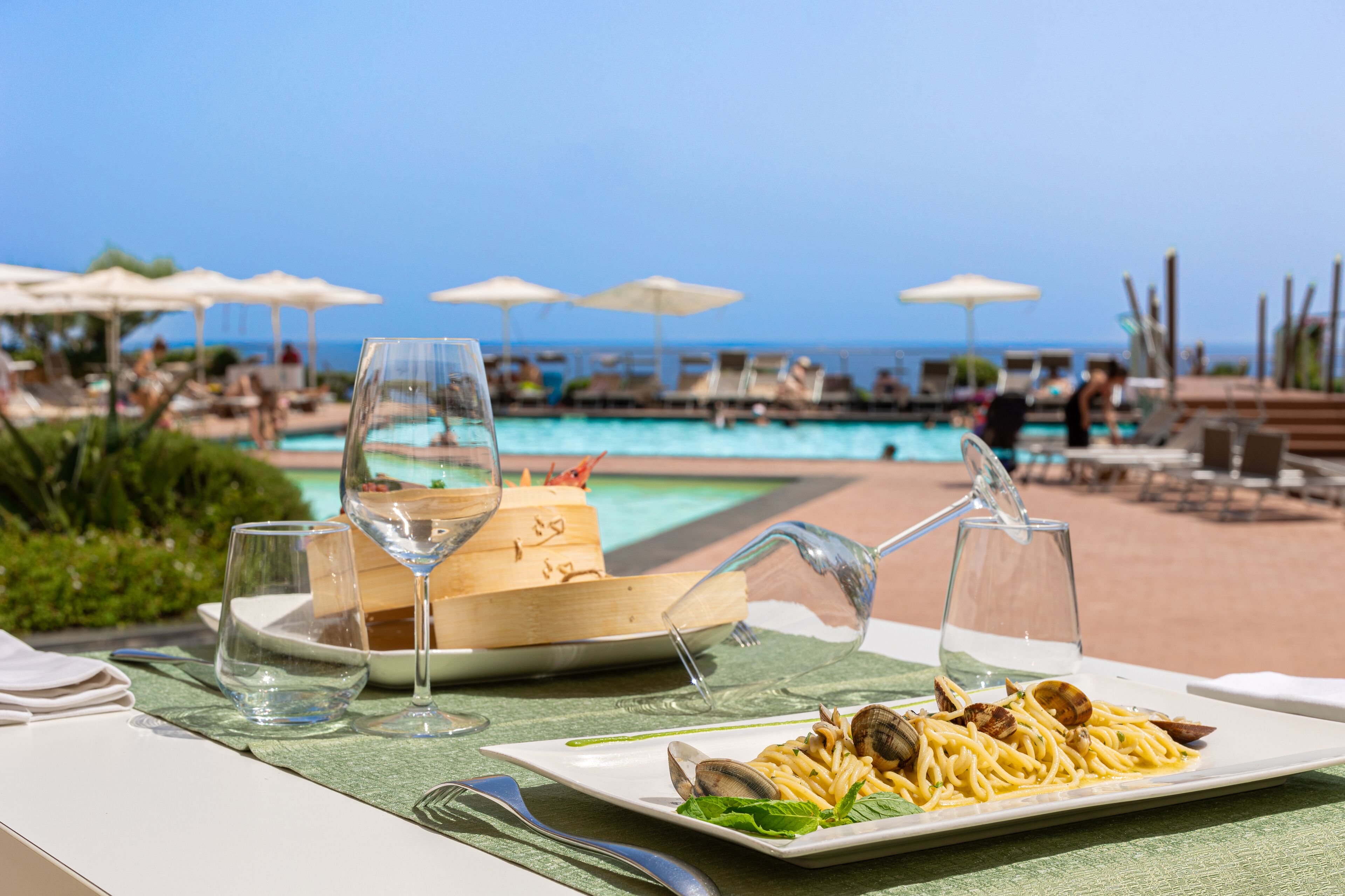 A dining table with a plate of pasta and clams, two wine glasses, and a menu on a green tablecloth by a poolside.