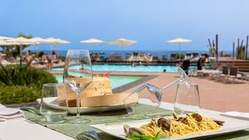 A dining table with a plate of pasta and clams, two wine glasses, and a menu on a green tablecloth by a poolside.