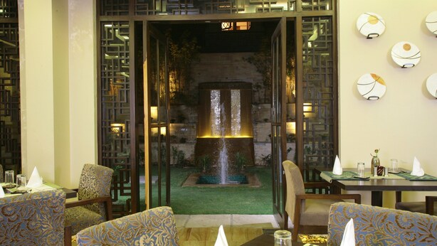 A dining area with tables set for two, chairs with patterned upholstery, a decorative wall with spherical light fixtures, and a view of a fountain through an open doorway. - Justa Gurgaon (Gurugram)