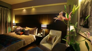 A hotel room with two beds, a sitting area with a chair, a nightstand, and a vase with flowers. - Justa Gurgaon (Gurugram)