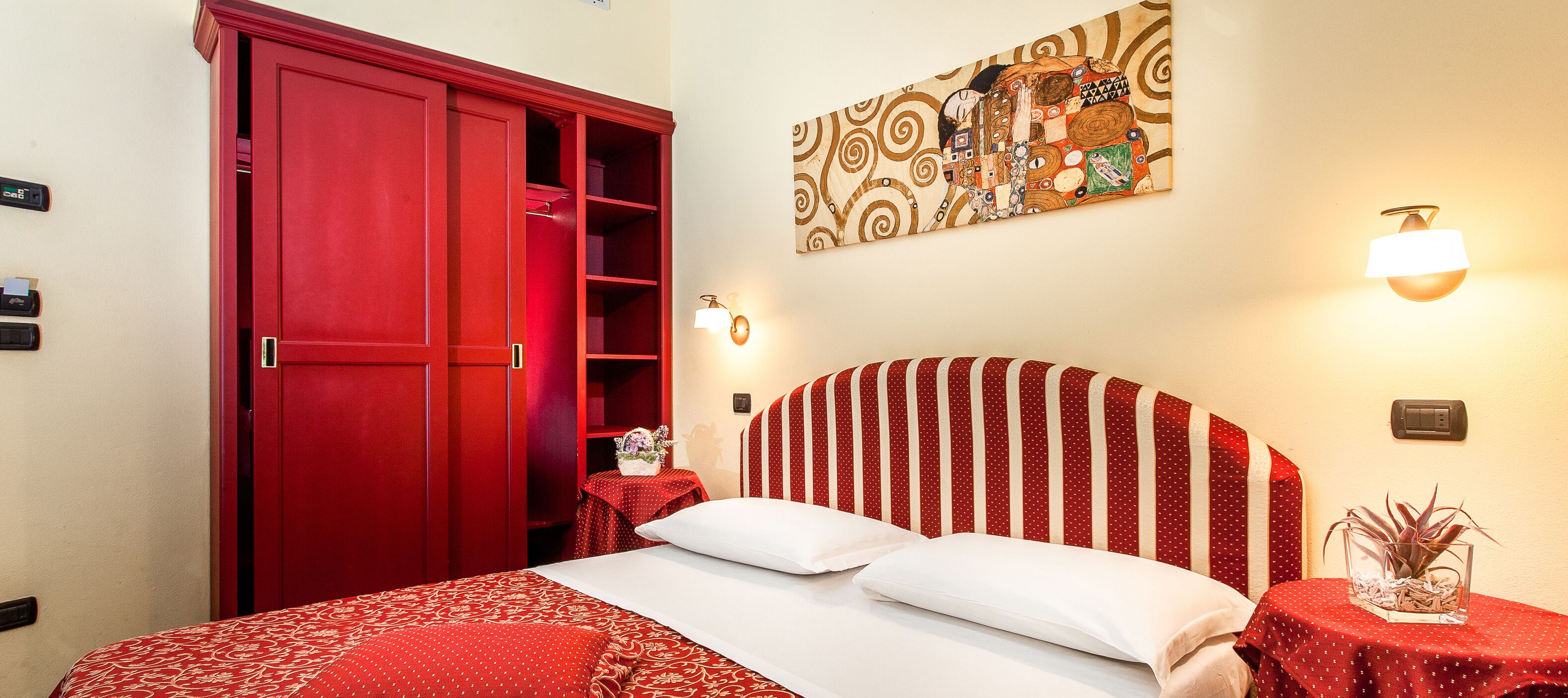 A hotel room with a red wardrobe, a bed with red and white bedding, a wall-mounted artwork, and a bedside table with a lamp.