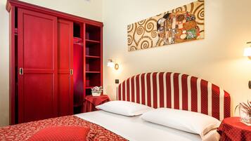 A hotel room with a red wardrobe, a bed with red and white bedding, a wall-mounted artwork, and a bedside table with a lamp.