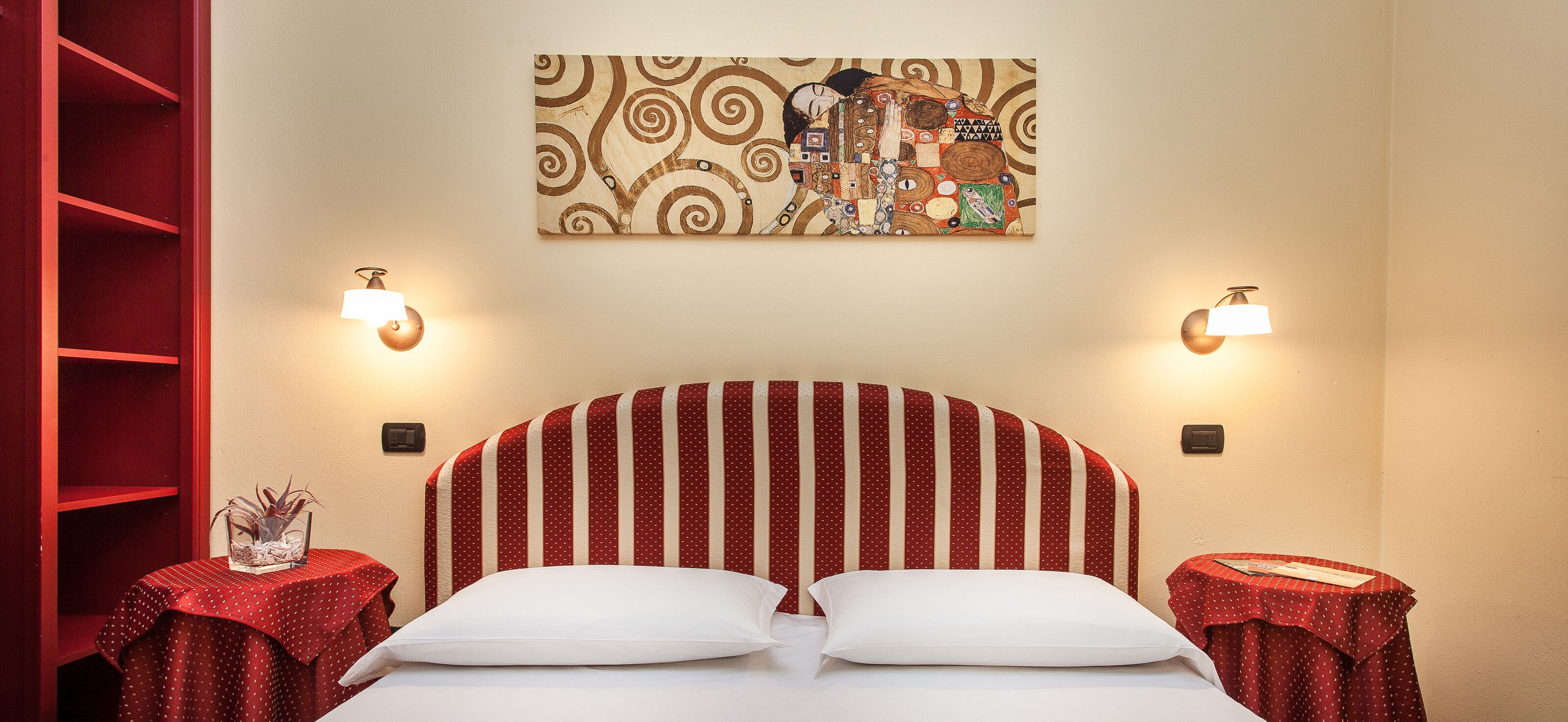 Une chambre d’hôtel avec une tête de lit à rayures, deux tables de chevet avec des lampes, une nappe à pois rouges et une œuvre d’art murale au-dessus du lit.